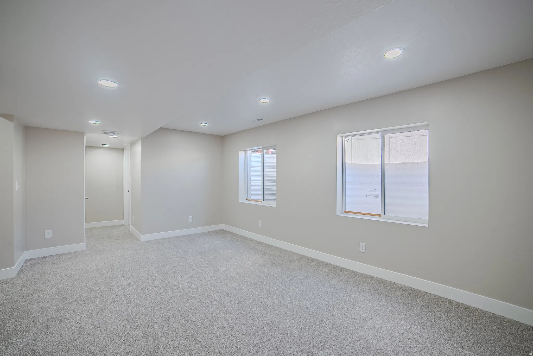 Spacious room featuring light gray carpet, recessed lighting, two windows with horizontal blinds, and clean white baseboards
