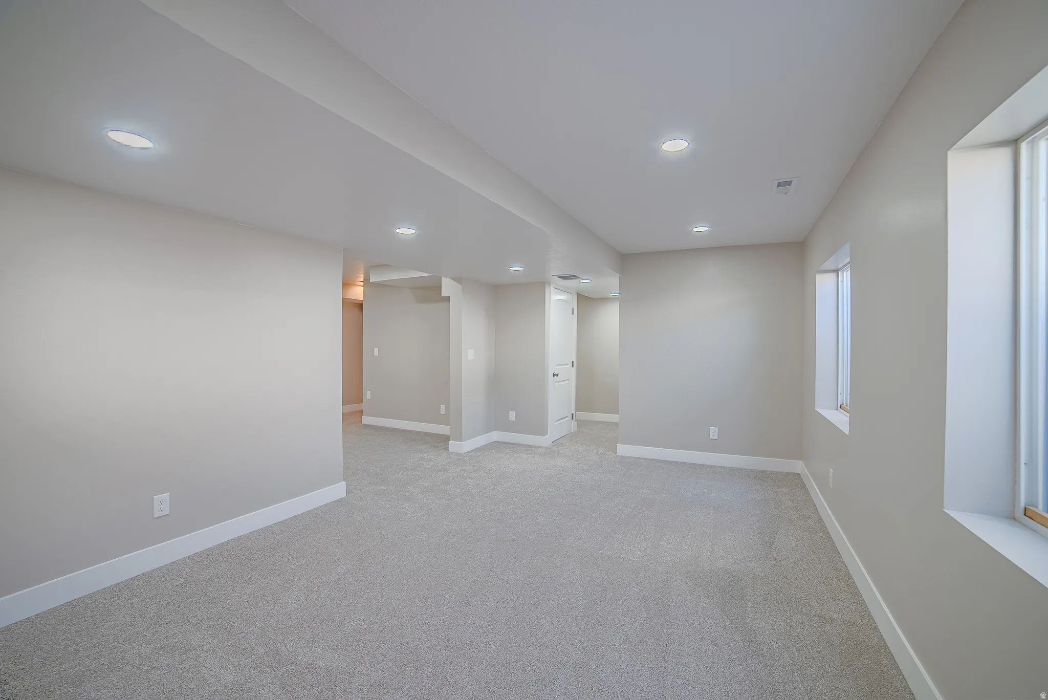 Spacious interior room featuring light-colored carpeting, recessed ceiling lighting, two windows, neutral wall tones, and white baseboards