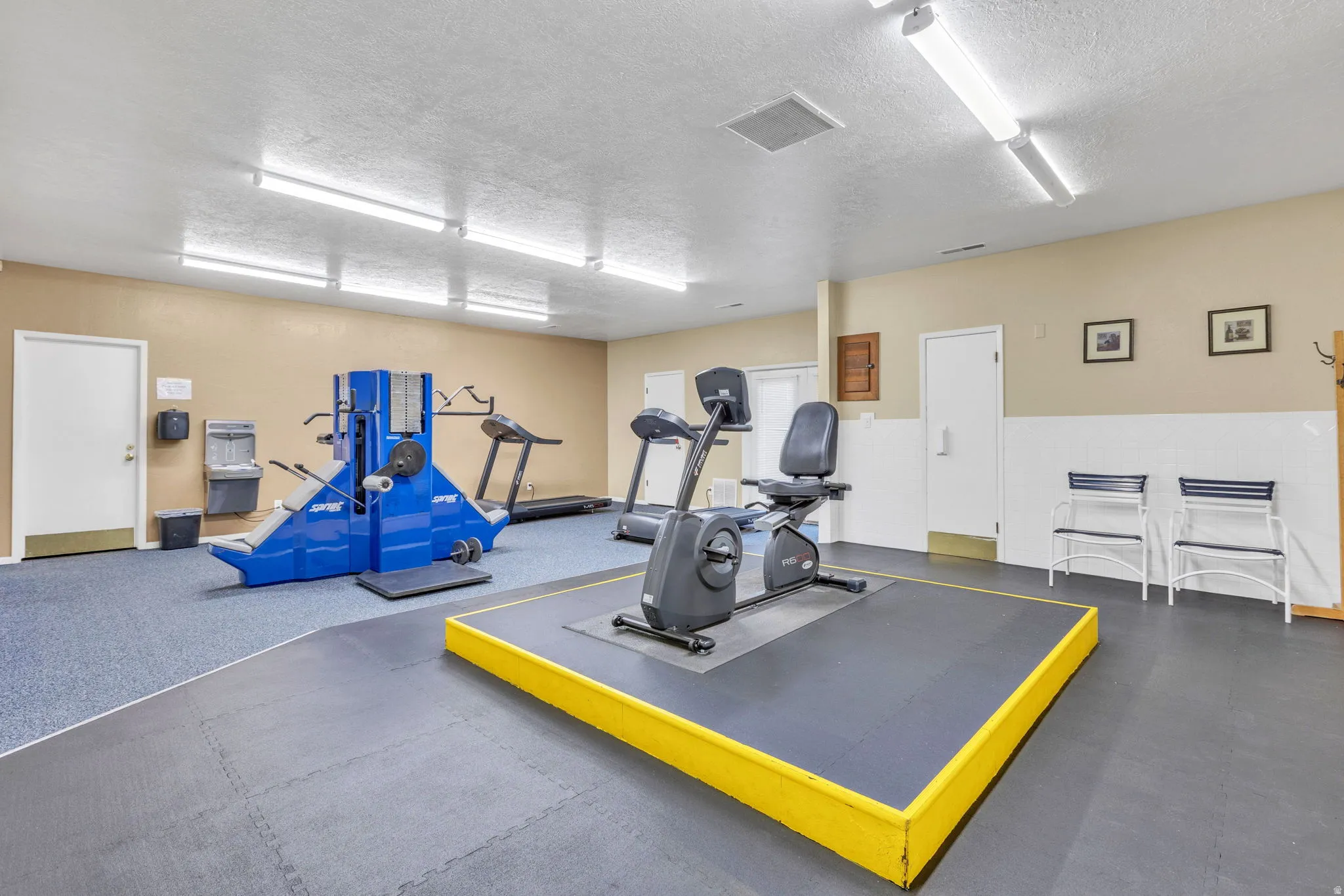 Dedicated fitness area featuring durable rubber flooring, a raised exercise platform, recessed ceiling lighting, and a beige and white wall palette