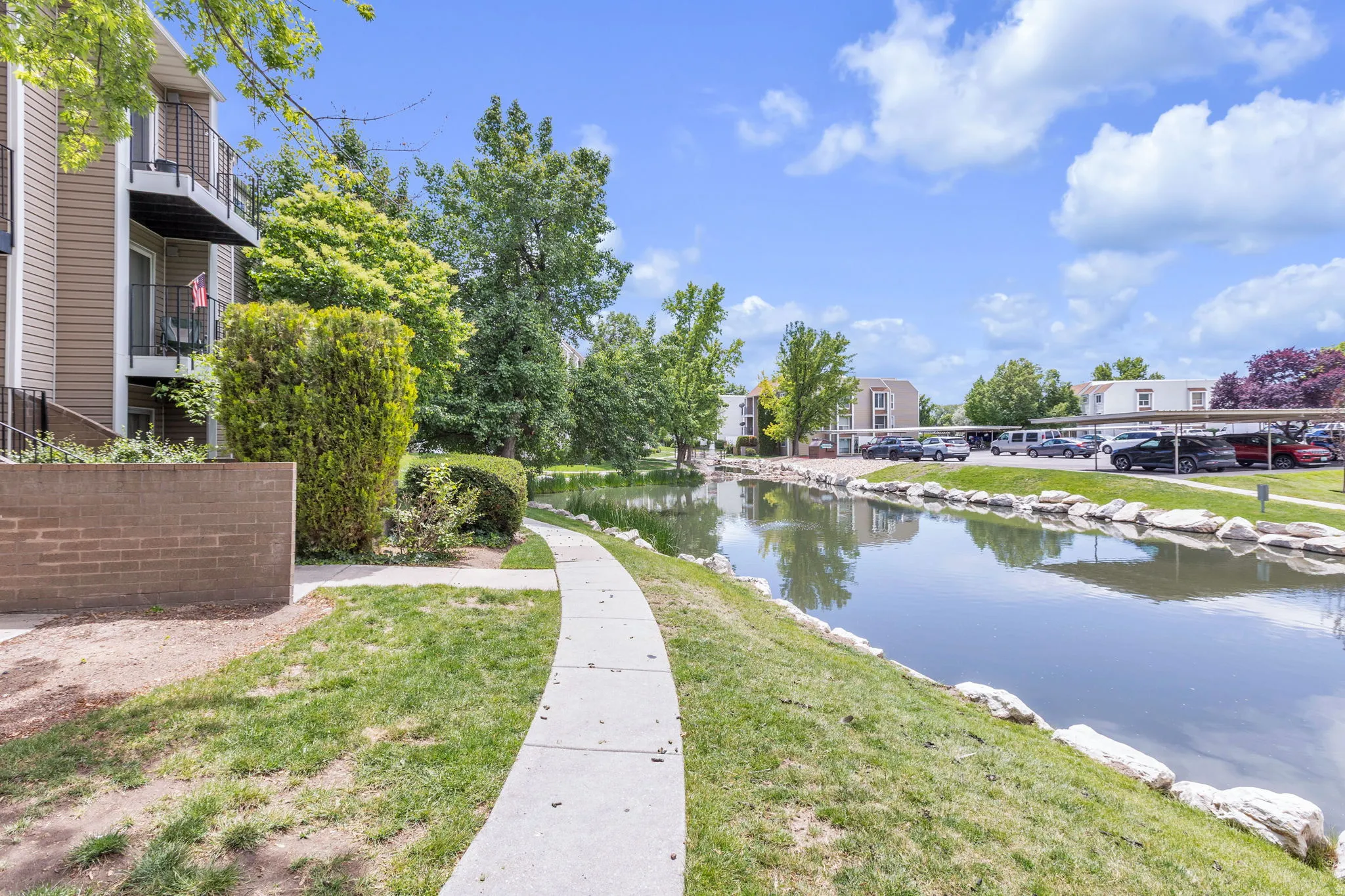 Manicured grounds featuring a paved pathway, a tranquil water feature, mature trees, and a building with private balconies