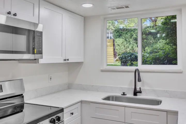 Kitchen featuring white shaker-style cabinetry, stainless steel appliances, a corner sink with a matte black faucet, light-toned countertops, and a window overlooking lush greenery