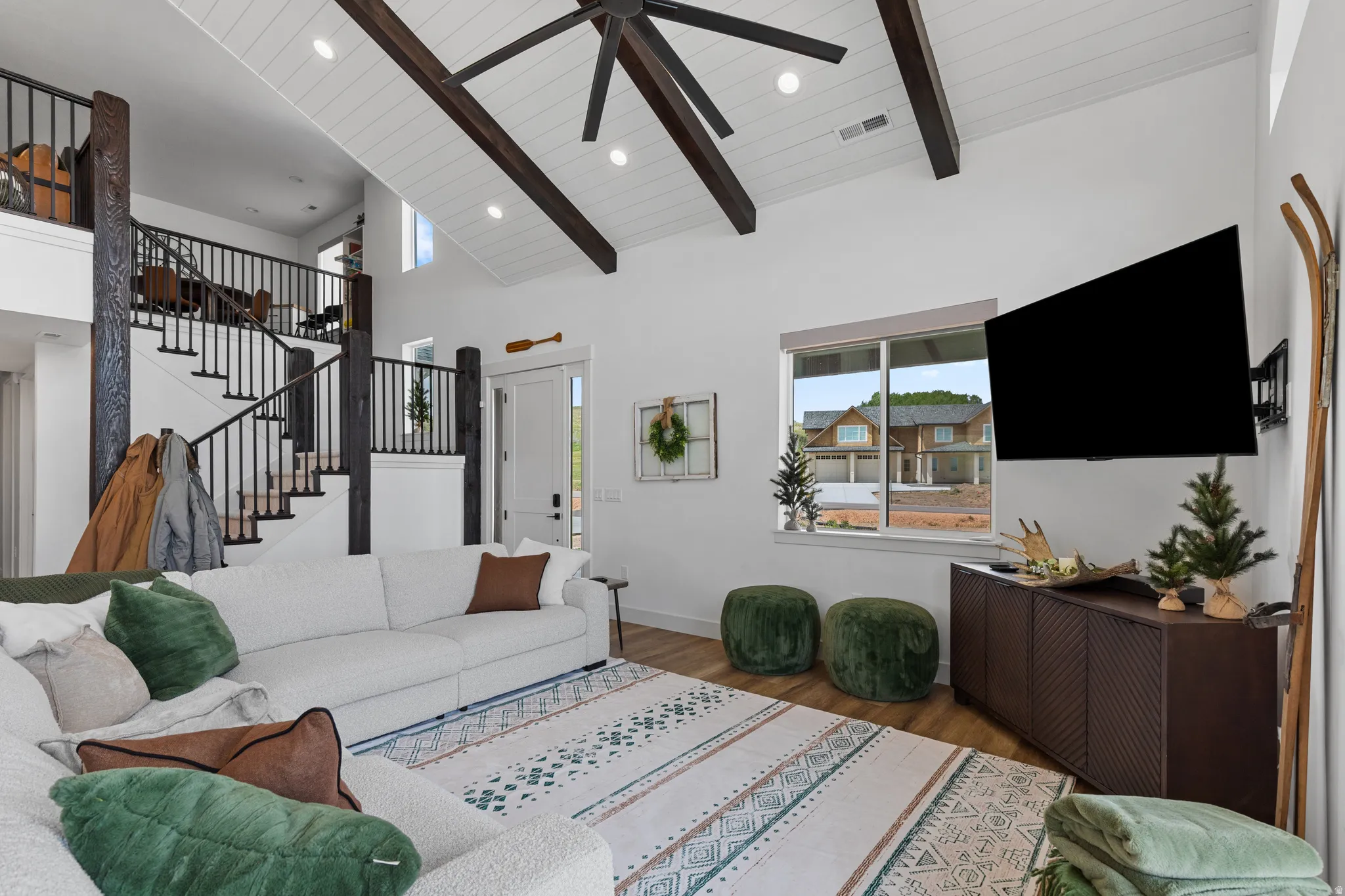 Spacious living area featuring a vaulted ceiling with exposed wood beams, wood-finish flooring, a contemporary ceiling fan, and large windows providing natural light