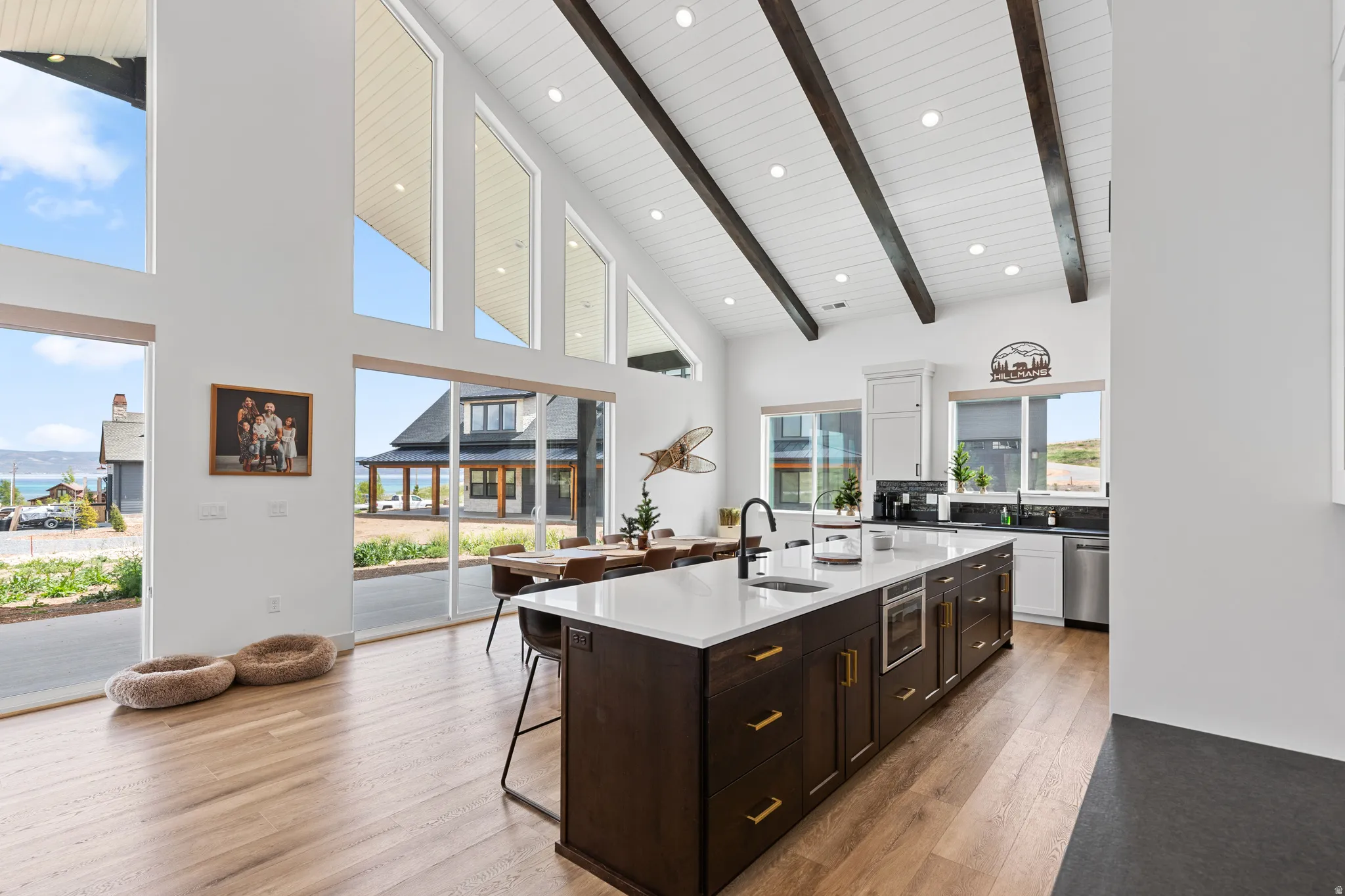 Open-concept great room featuring a vaulted ceiling with exposed wood beams, expansive windows, light wood-finish flooring, a contemporary kitchen island with a white countertop, and integrated stainless steel appliances