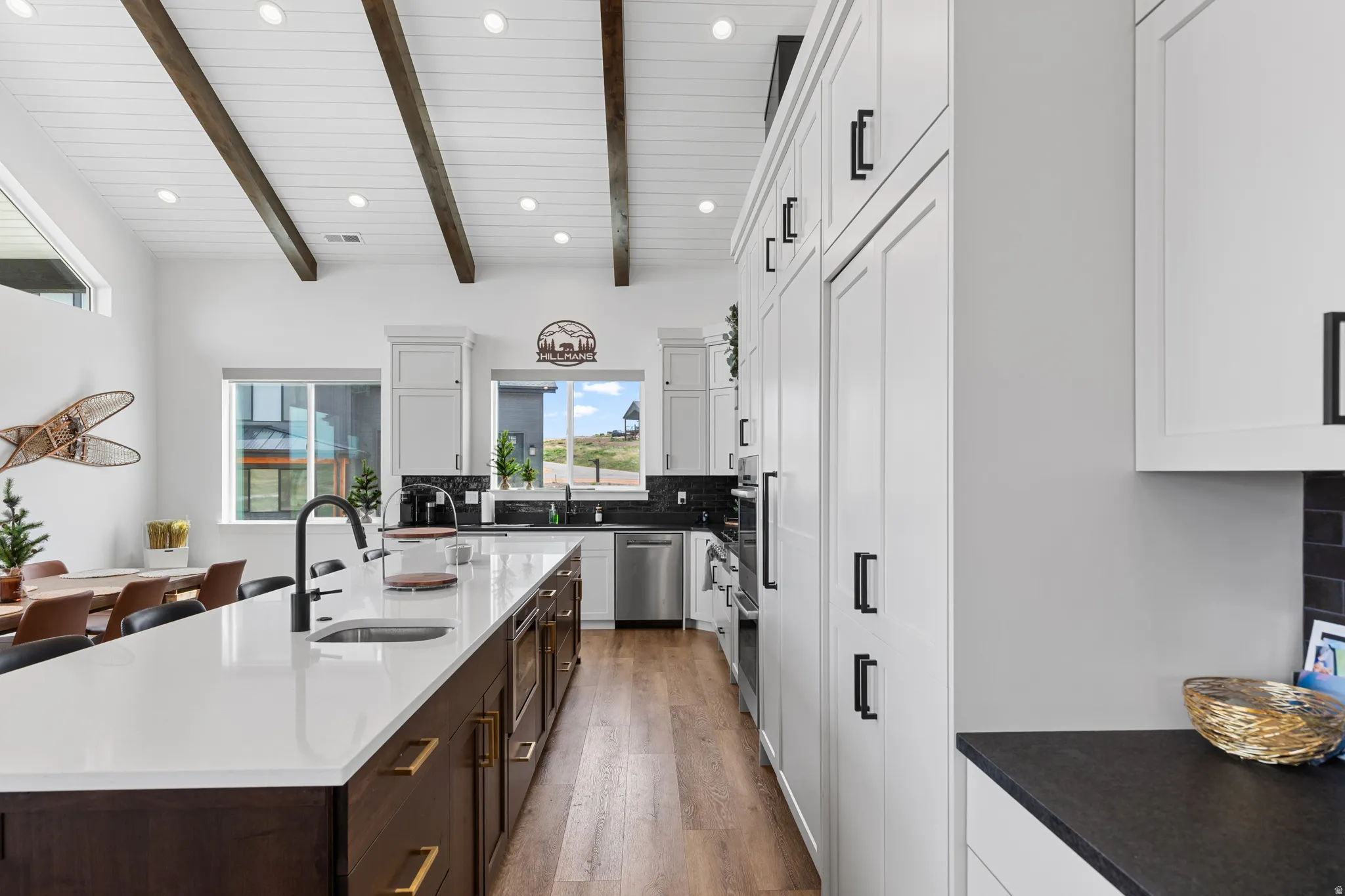 Spacious kitchen featuring a large central island with a white countertop and dark wood-finish cabinetry, stainless steel appliances, white flat-panel cabinetry with black hardware, and wood-finish flooring