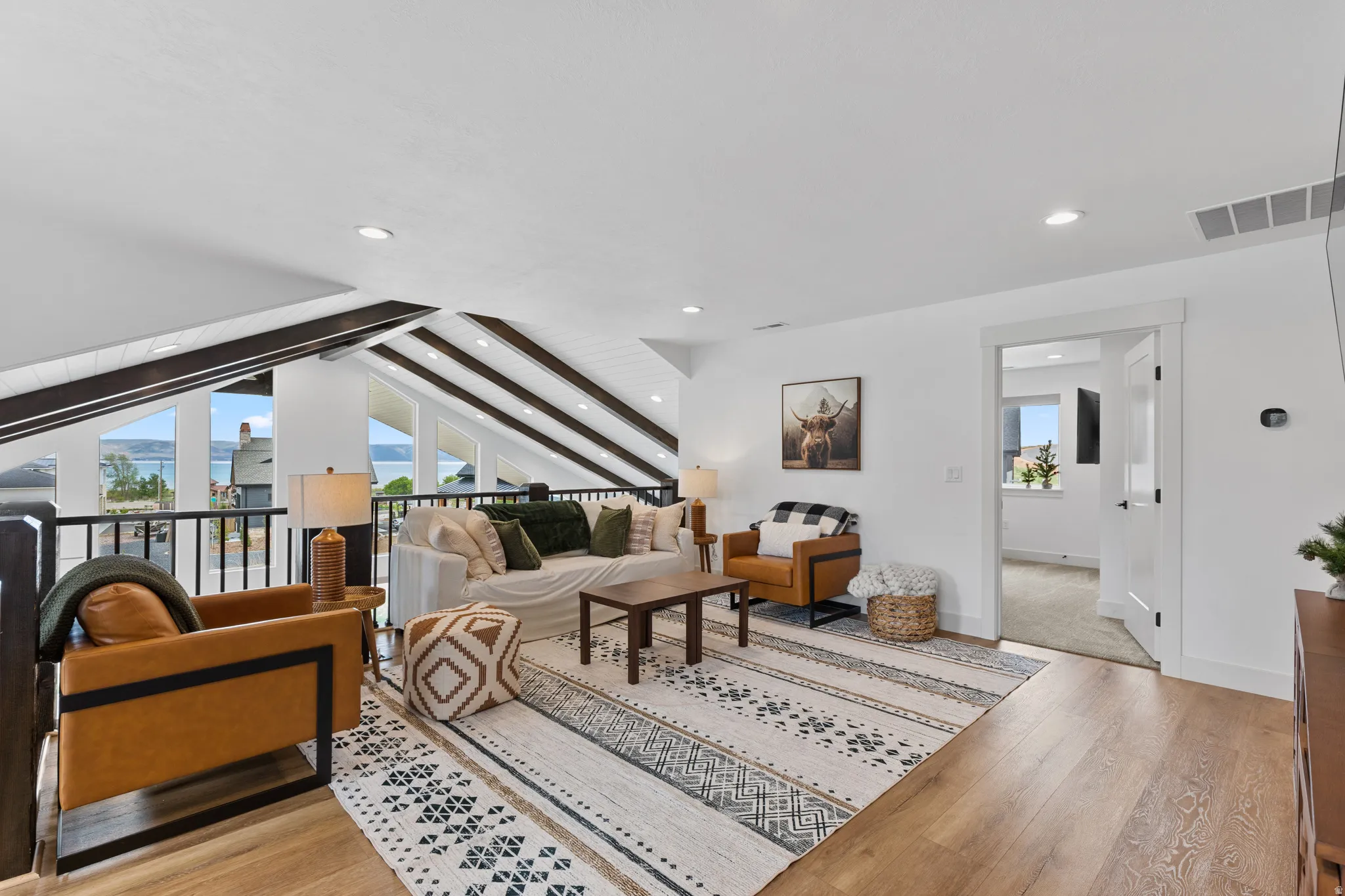 Spacious loft area featuring wood-finish flooring, a vaulted ceiling with exposed beams, recessed lighting, and a white interior door leading to an additional room