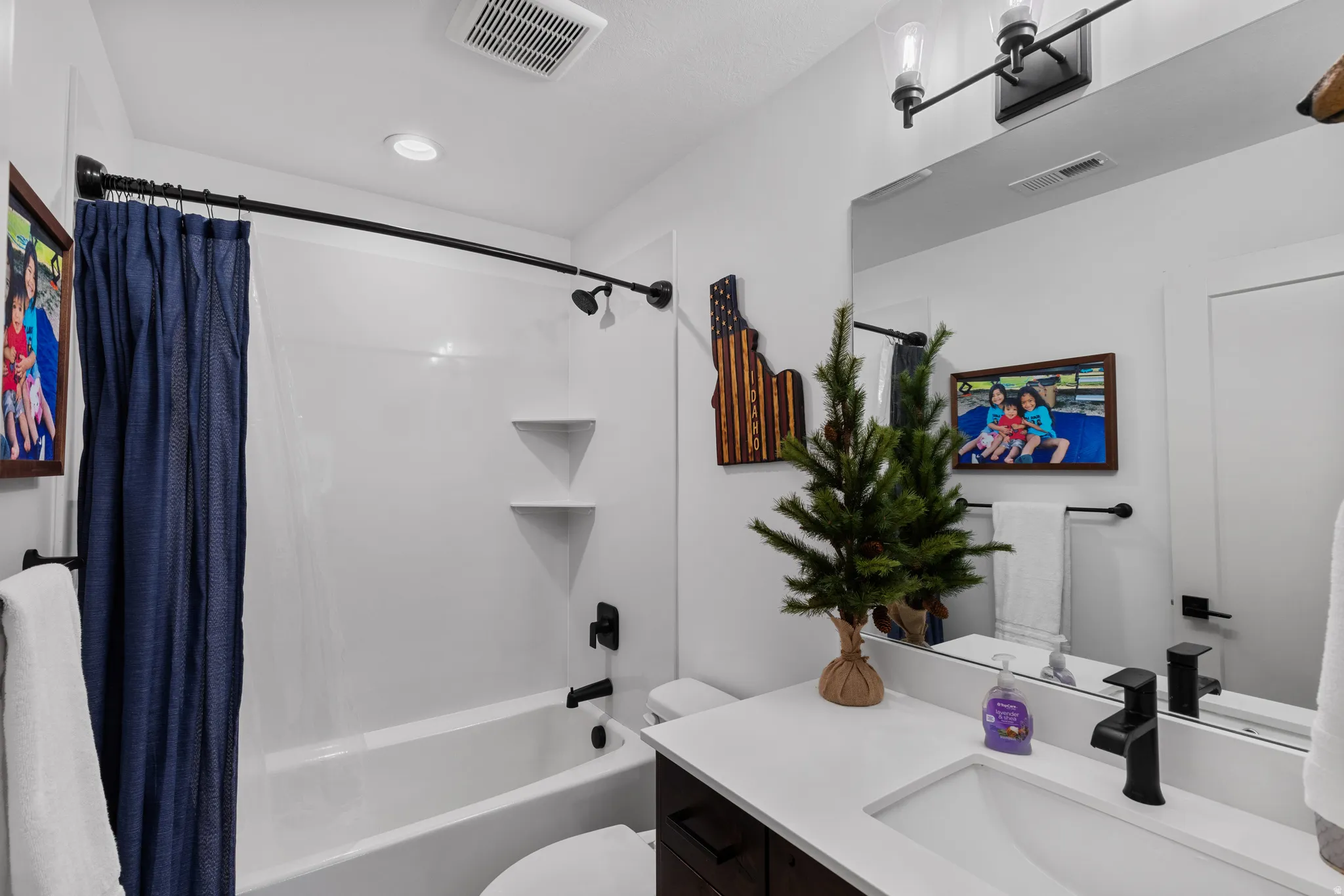 Bathroom featuring a white bathtub and shower combination with black fixtures, a single vanity with a white countertop and integrated sink, a large wall-mounted mirror, and a modern two-light fixture