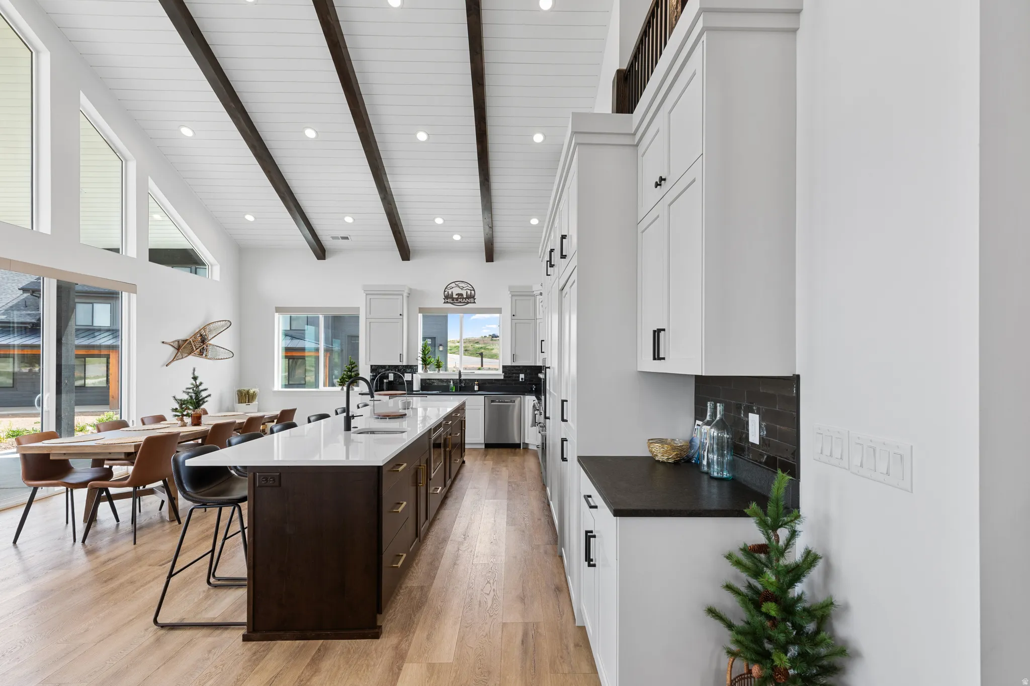 Open-concept living area featuring a vaulted ceiling with exposed dark wood beams, light wood-finish flooring, and a spacious kitchen island with a white countertop