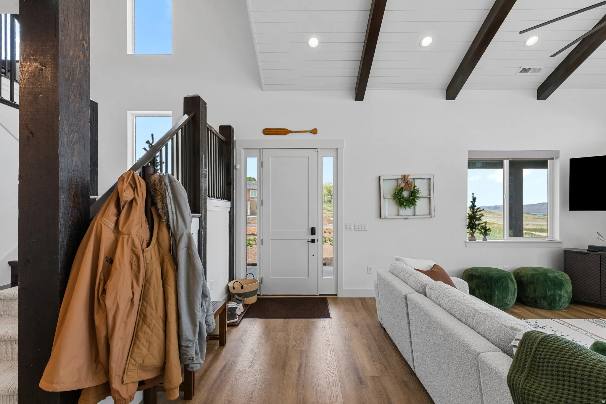 Vaulted ceiling with exposed wood beams, wood-finish flooring, white paneled entry door with sidelights, and large windows providing natural light
