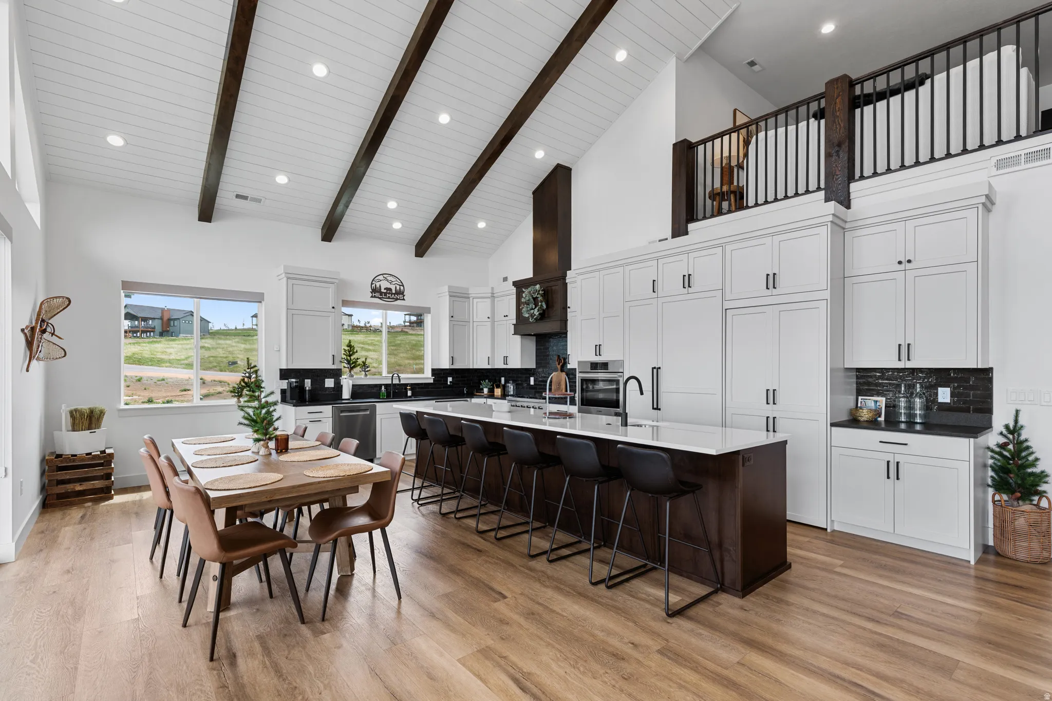 Spacious kitchen and dining area featuring a vaulted ceiling with exposed wood beams, wood-finish flooring, white cabinetry, an oversized island with seating, and a built-in pantry system