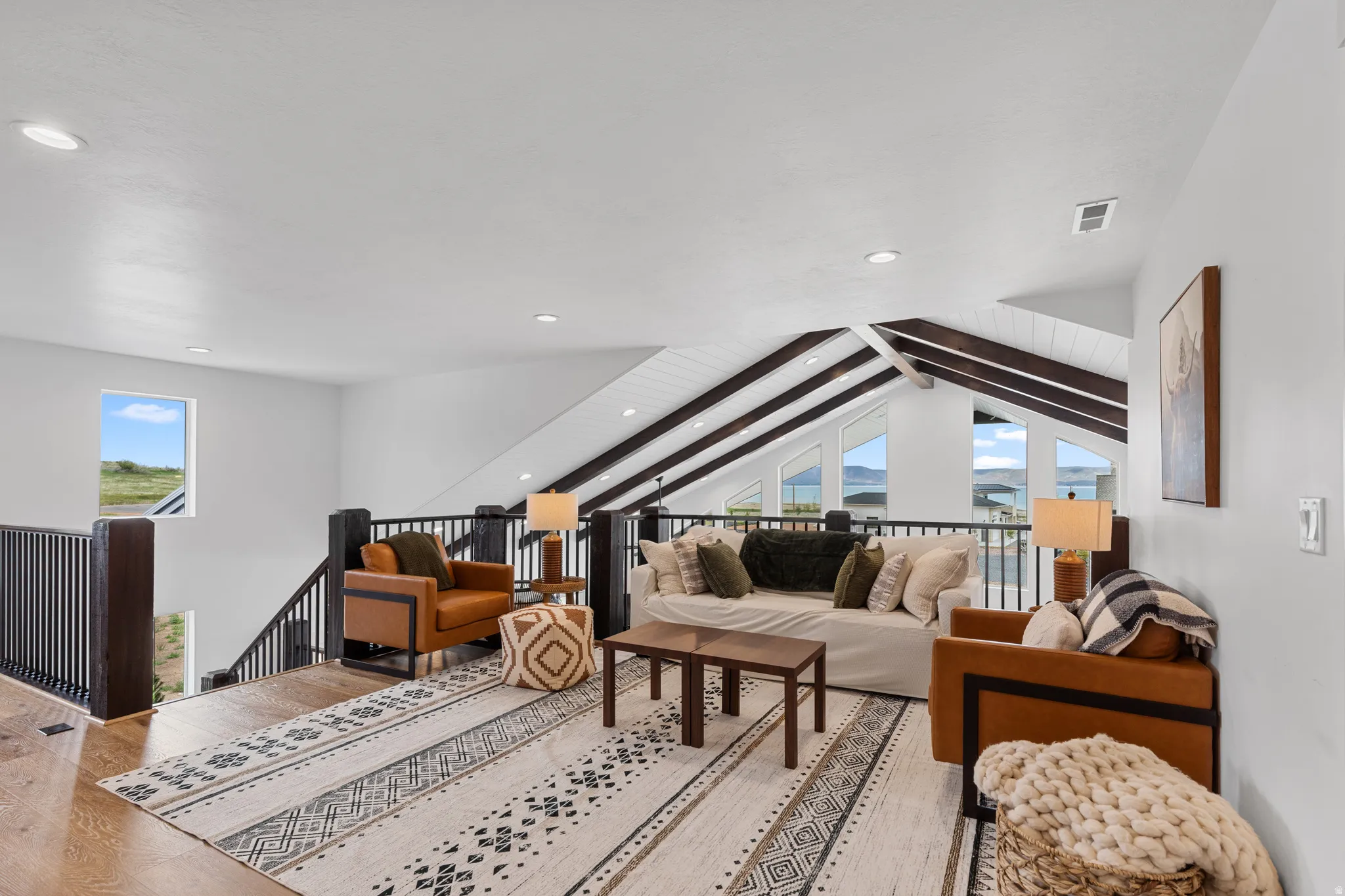 Bright open loft space featuring vaulted ceilings with exposed beams, wood-finish flooring, recessed lighting, and a dark wood and metal railing system