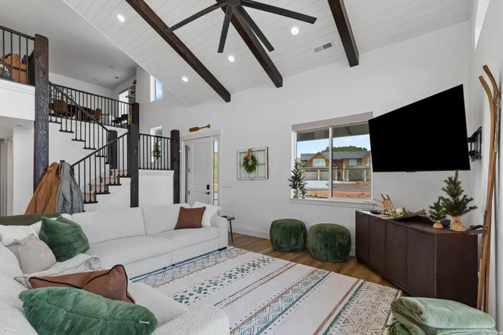 Spacious living area featuring a vaulted ceiling with exposed wood beams, wood-finish flooring, a contemporary ceiling fan, and large windows providing natural light