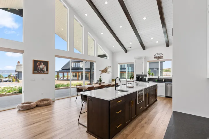 Open-concept great room featuring a vaulted ceiling with exposed wood beams, expansive windows, light wood-finish flooring, a contemporary kitchen island with a white countertop, and integrated stainless steel appliances