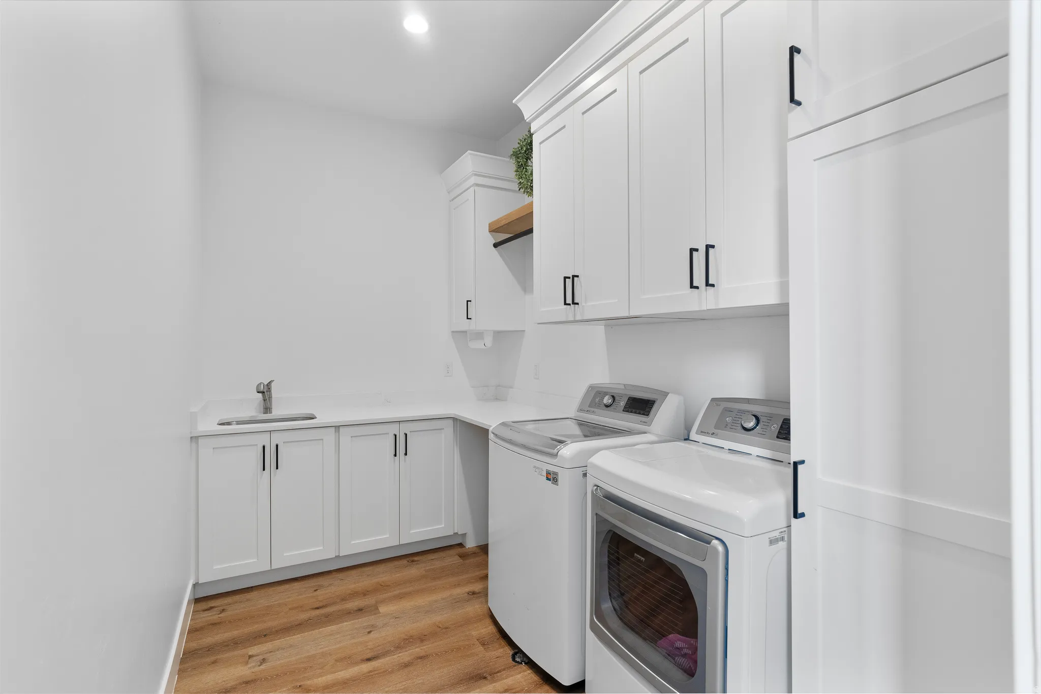 Dedicated laundry room featuring wood-finish flooring, white cabinetry with matte black hardware, white countertops, and an integrated utility sink with a stainless steel faucet.