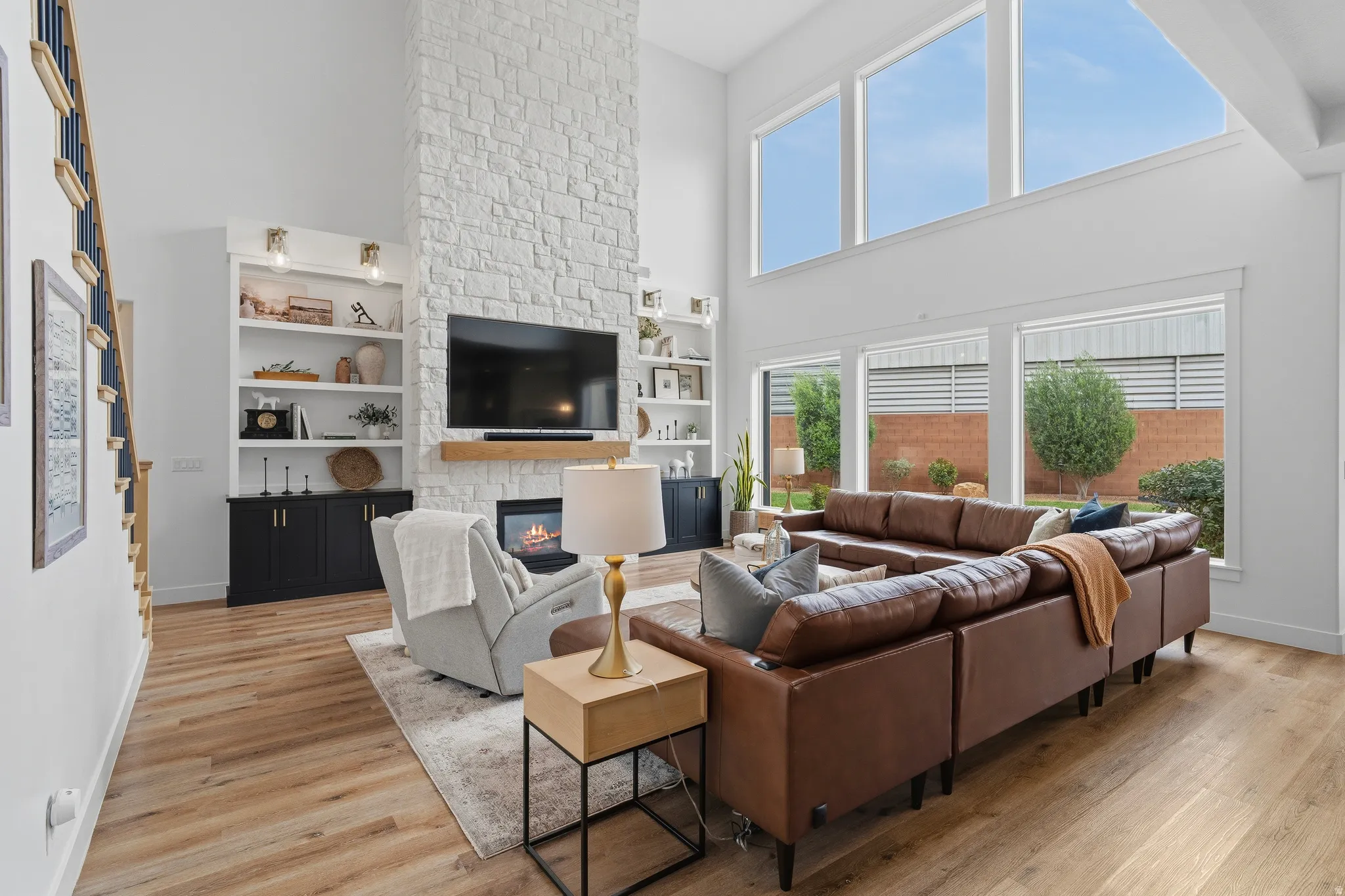 Vaulted living space featuring a stacked stone fireplace with wood mantel, large windows, wood-finish flooring, and built-in shelving with lower cabinetry.