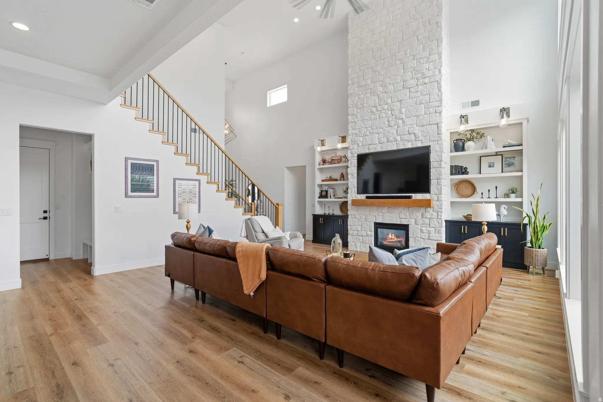 Vaulted ceiling living area featuring a floor-to-ceiling stone-clad fireplace with wood mantel, built-in shelving, wood-finish flooring, and a staircase with black metal balusters.