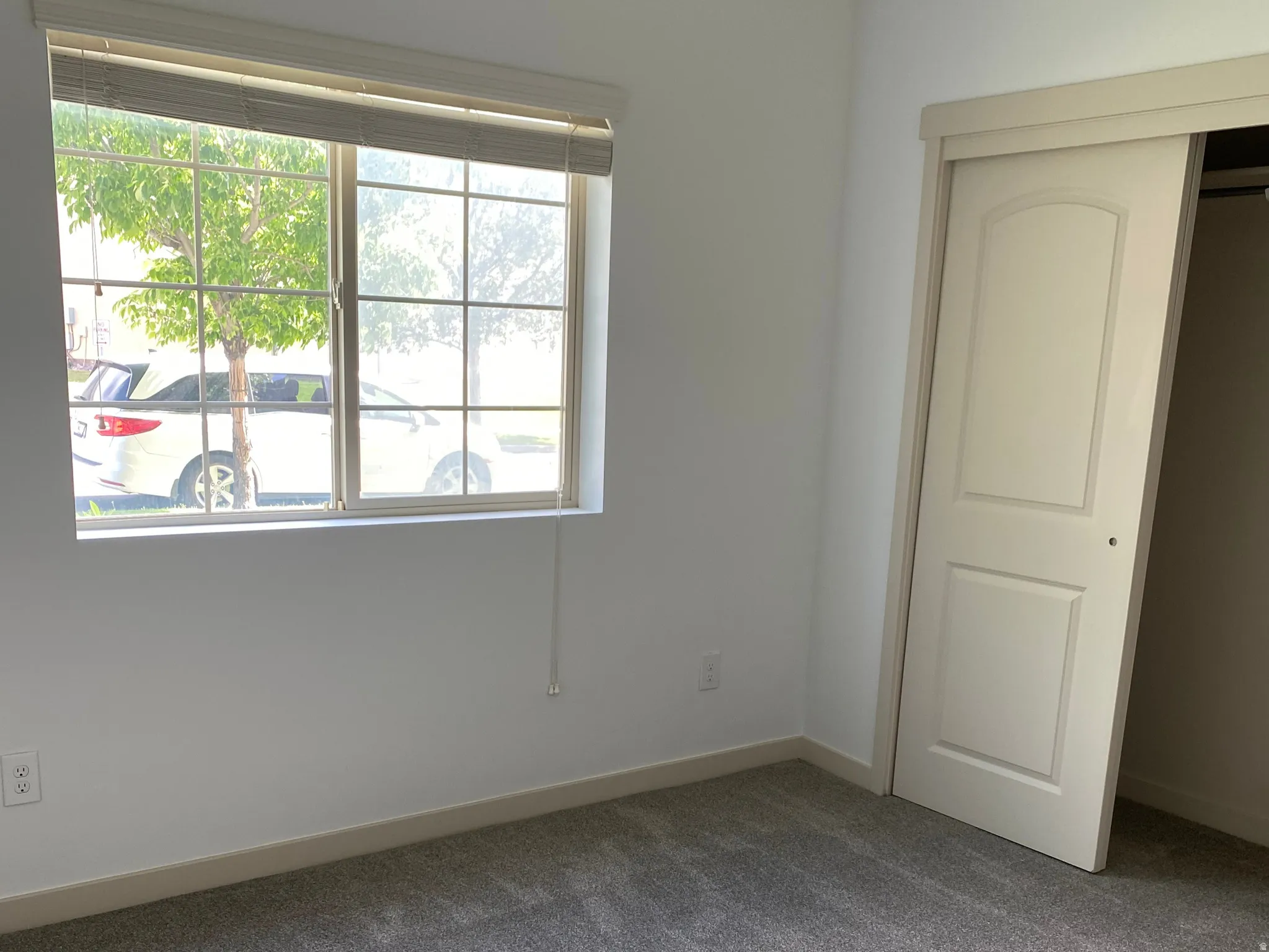 Carpeted interior space featuring a window with blinds, white walls, and a bi-fold closet door