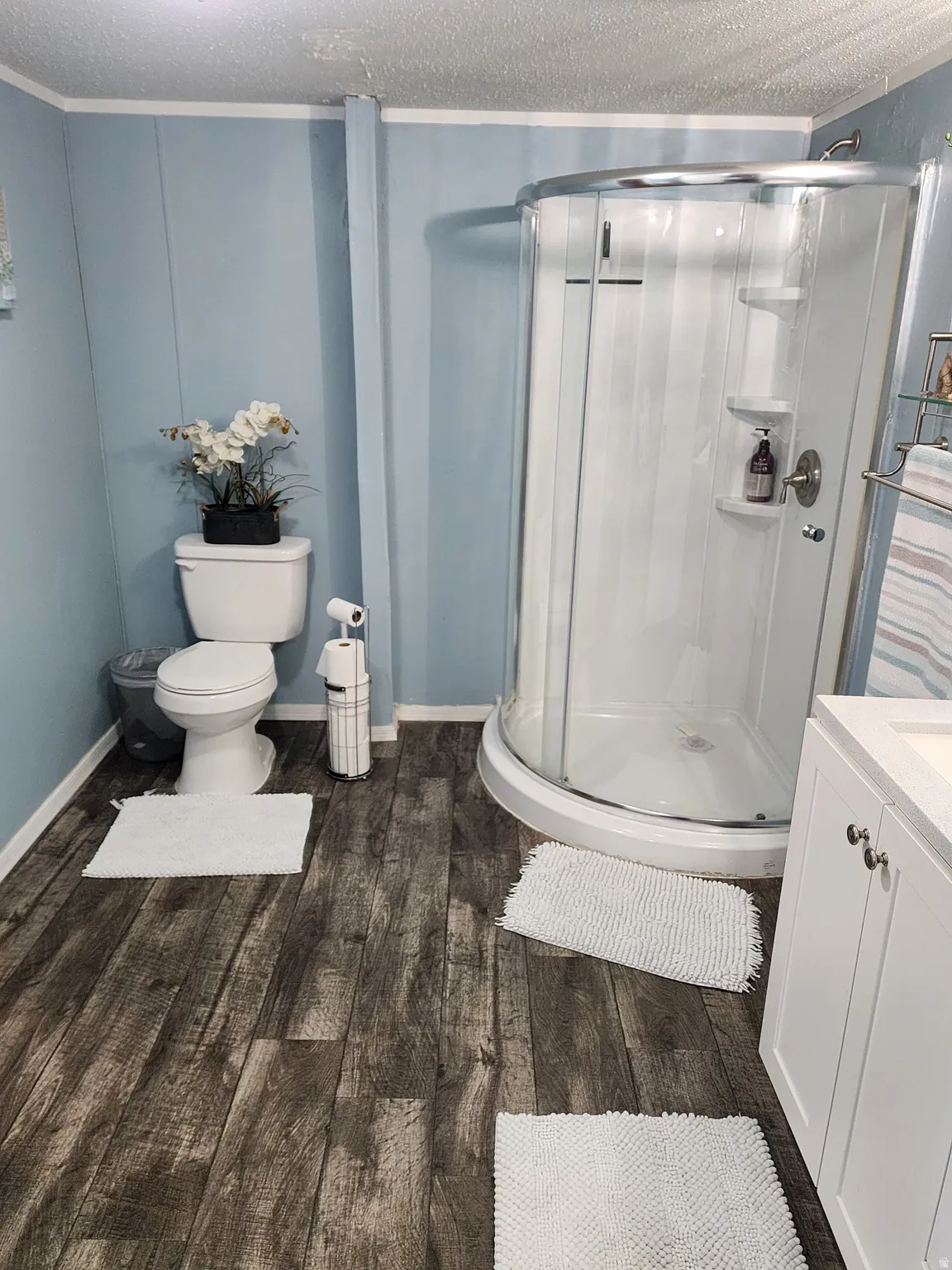 Three-piece bathroom featuring a corner shower enclosure, light blue wall panels, and wood-finish flooring