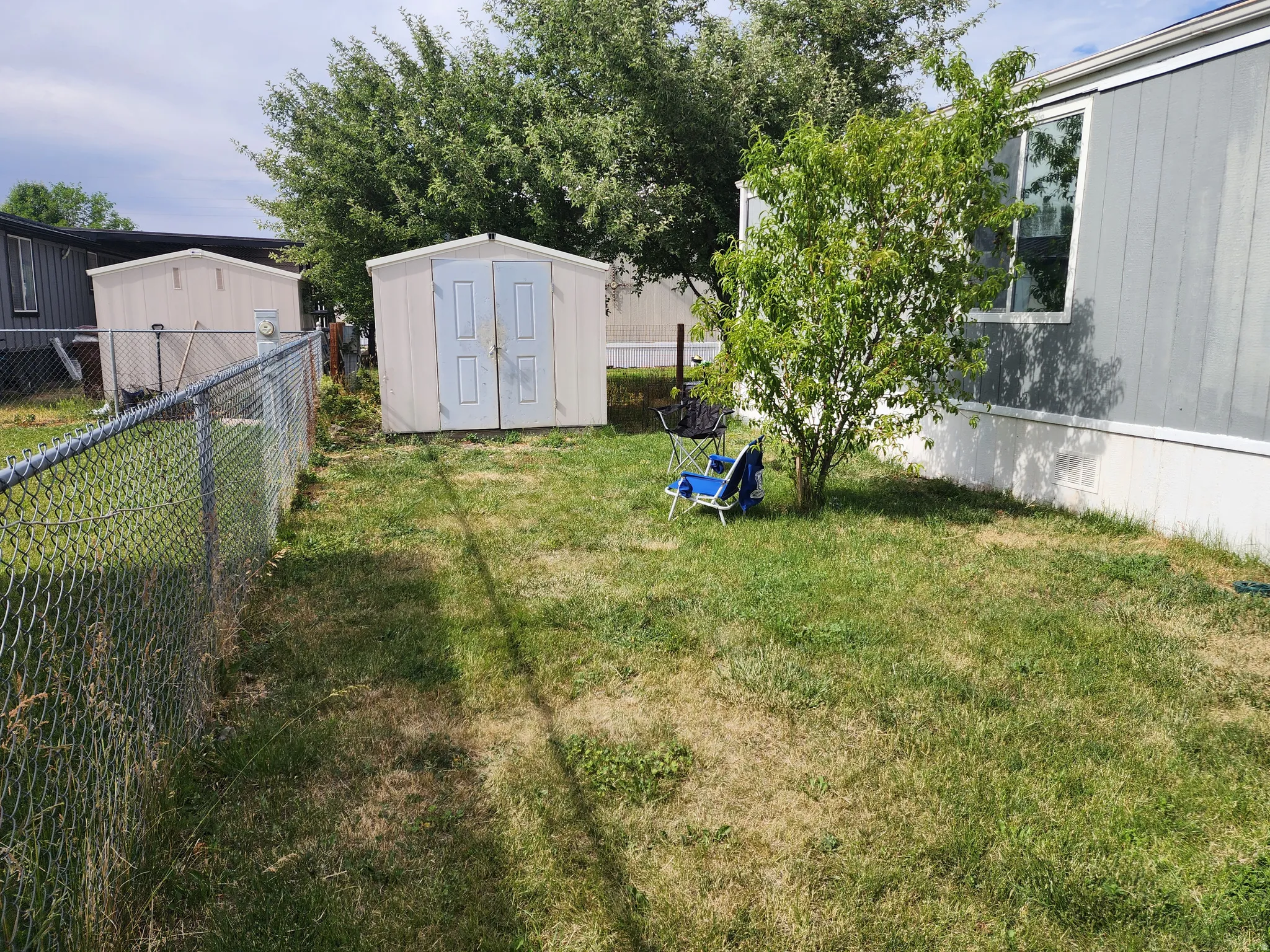 Fenced yard area featuring two storage sheds, a mature shrub, and a light-colored mobile home exterior with multiple windows