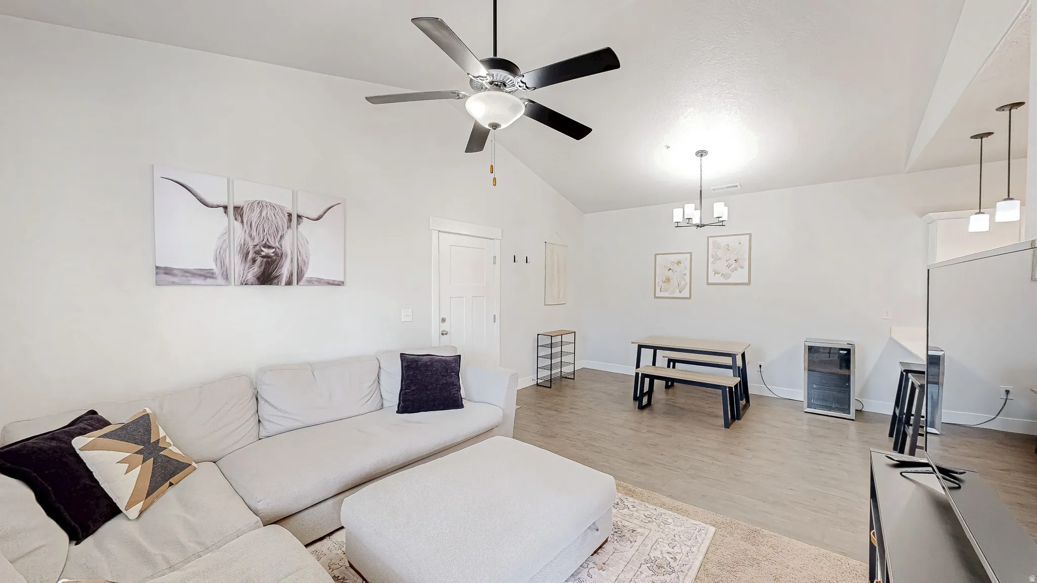 Open-concept living space featuring a vaulted ceiling with a ceiling fan, light wood-finish flooring, a dining area with a modern chandelier, and a breakfast bar with pendant lighting