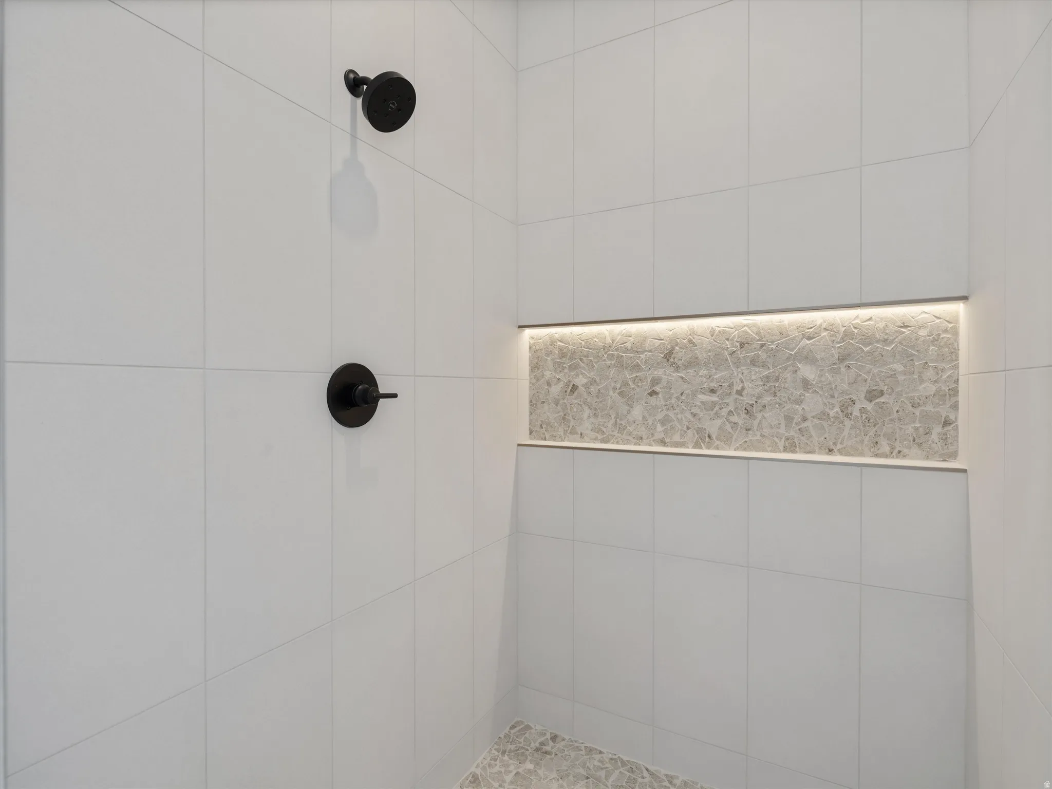 Shower stall featuring matte black fixtures, white tile surround, illuminated niche with stone accent, and coordinating stone shower pan