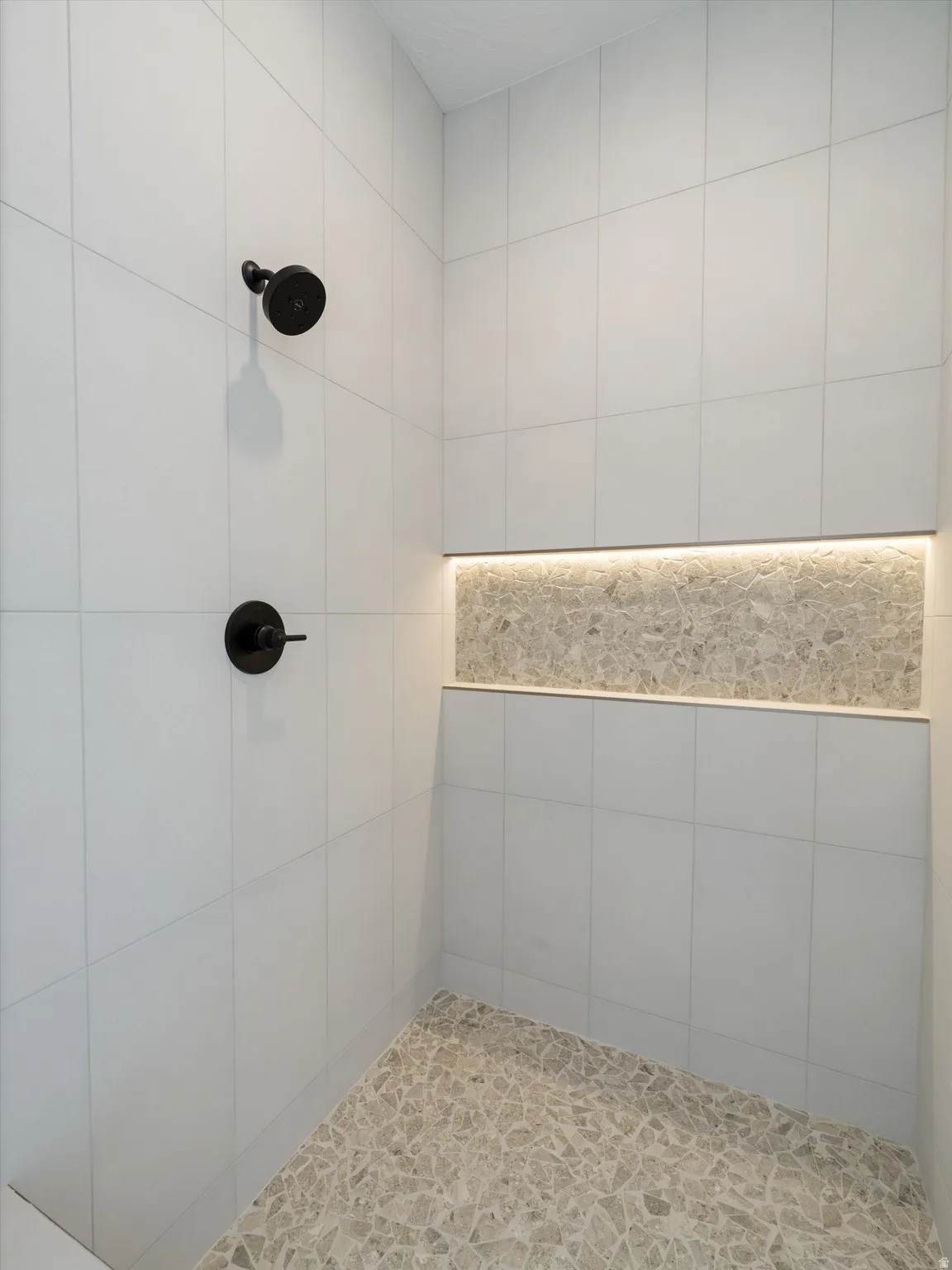 Shower with white rectangular wall tiles, matte black shower fixtures, mosaic tile flooring, and an illuminated niche with mosaic backing