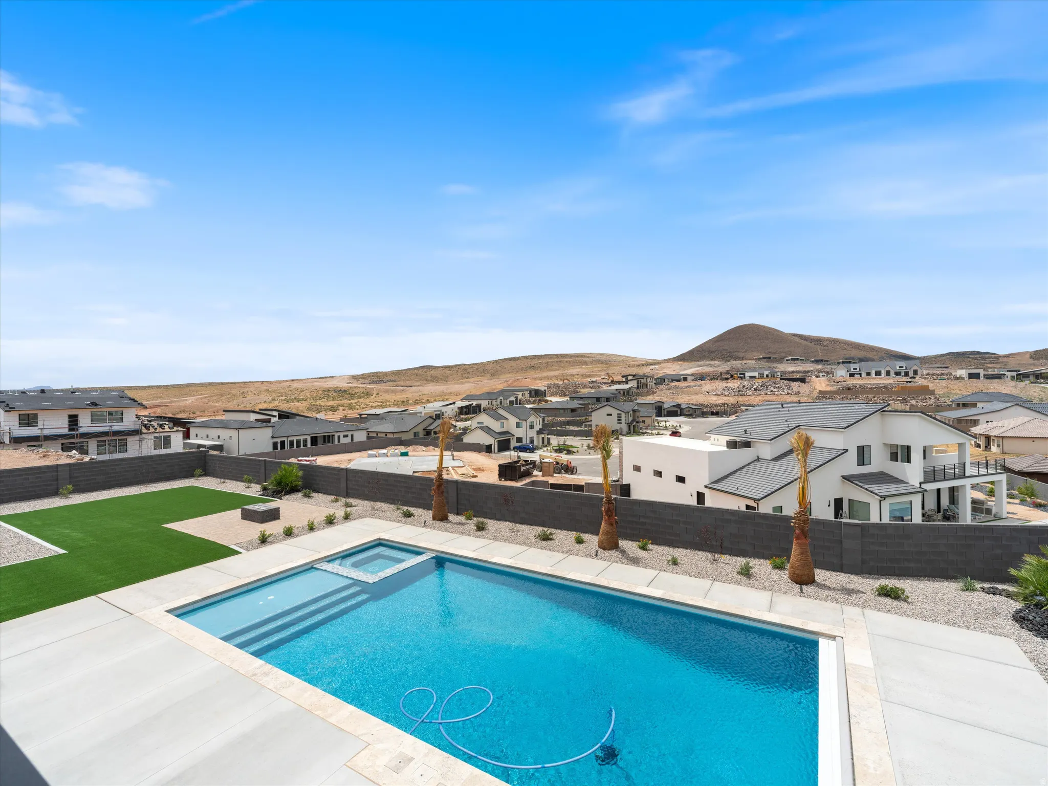 Resort-style backyard featuring a rectangular swimming pool with integrated spa, broad concrete decking, artificial turf lawn, and a low-maintenance gravel landscape with accent palms and a privacy fence