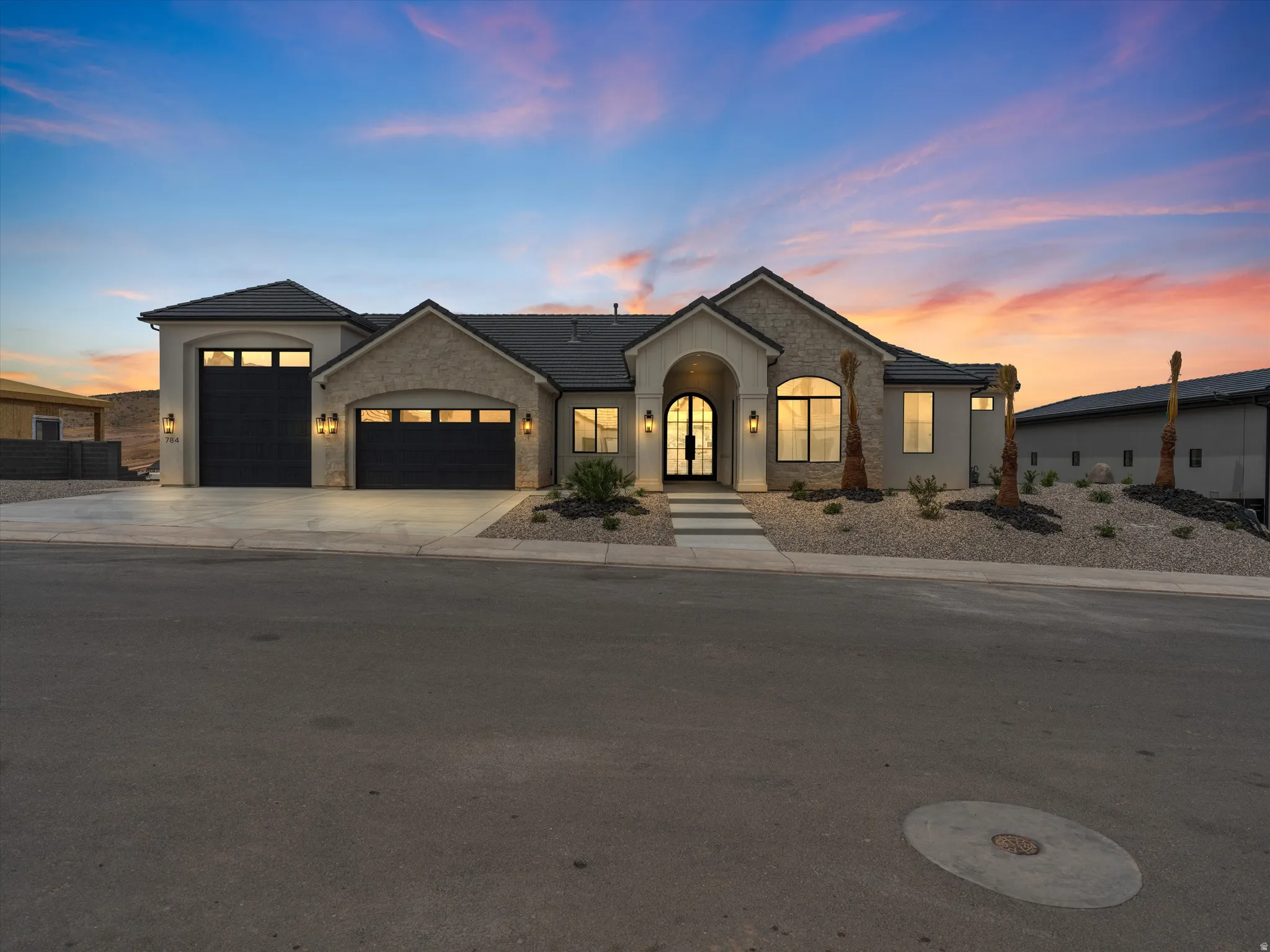 Contemporary stucco and stone exterior featuring an arched entryway with double glass doors, a multi-car garage with black doors and window inserts, exterior sconce lighting, and a xeriscaped front yard