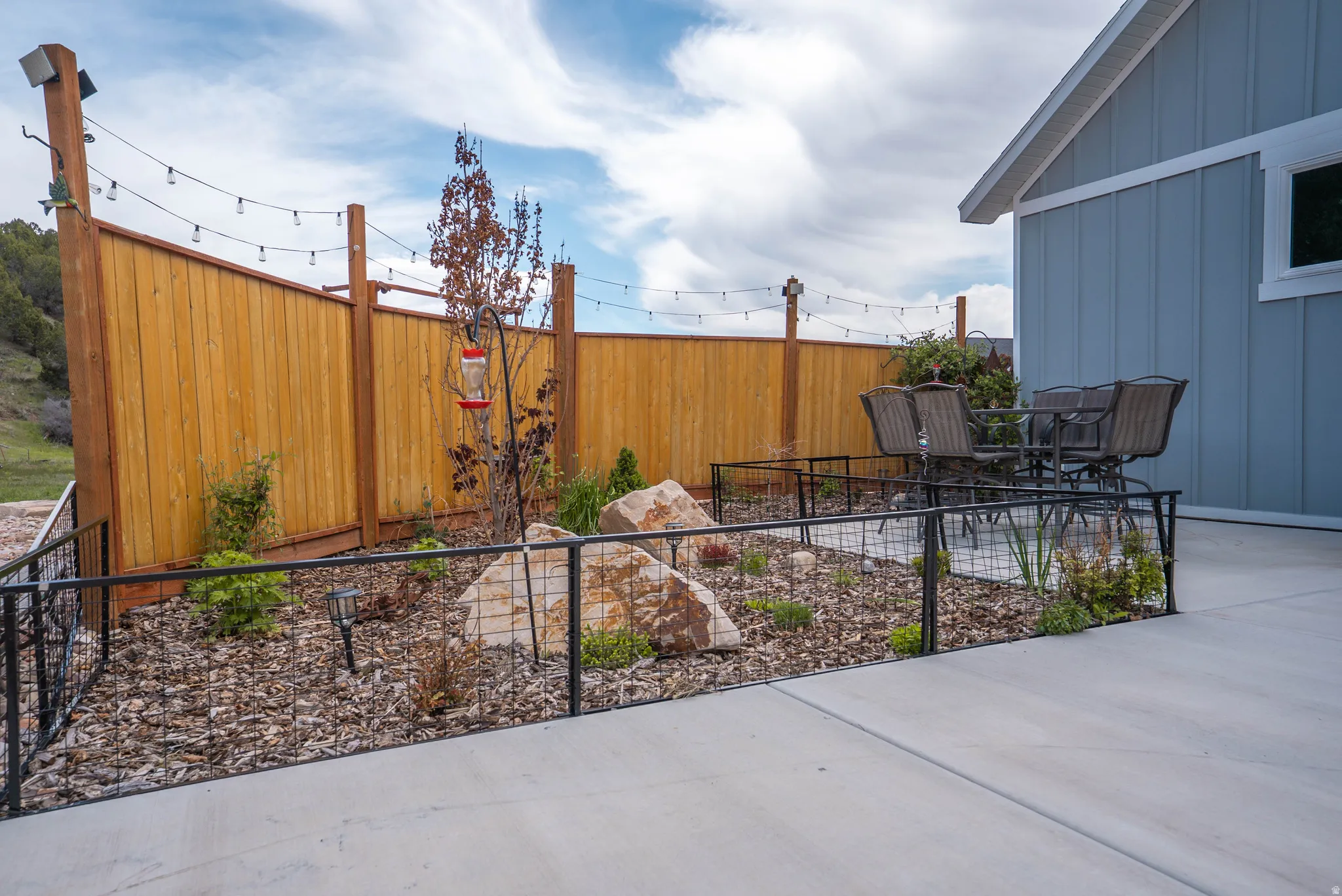 Concrete patio with natural stone landscaping featuring a privacy fence, string lighting, and board and batten siding