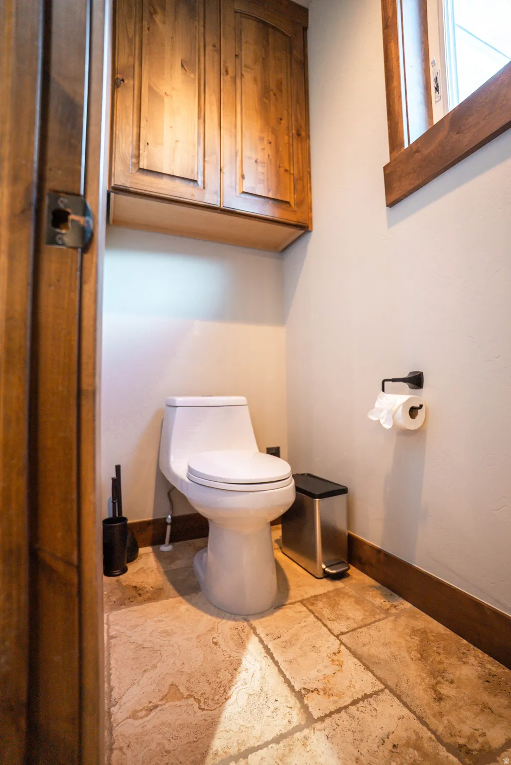 Compact water closet featuring natural wood cabinetry, wood-framed window, and stone tile flooring