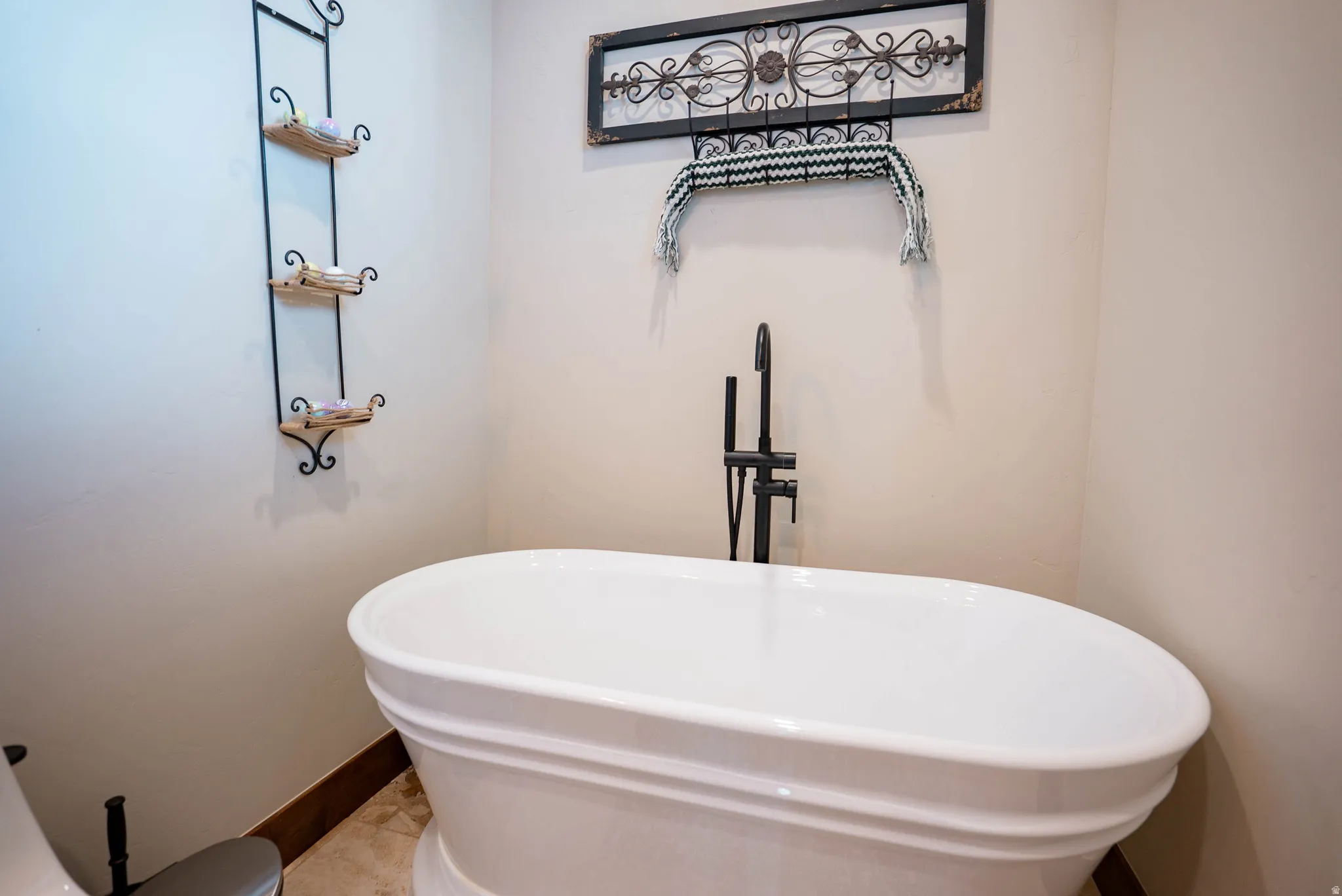 Freestanding soaking tub featuring a matte black floor-mounted tub filler, light-toned walls, and tile flooring with a dark wood baseboard