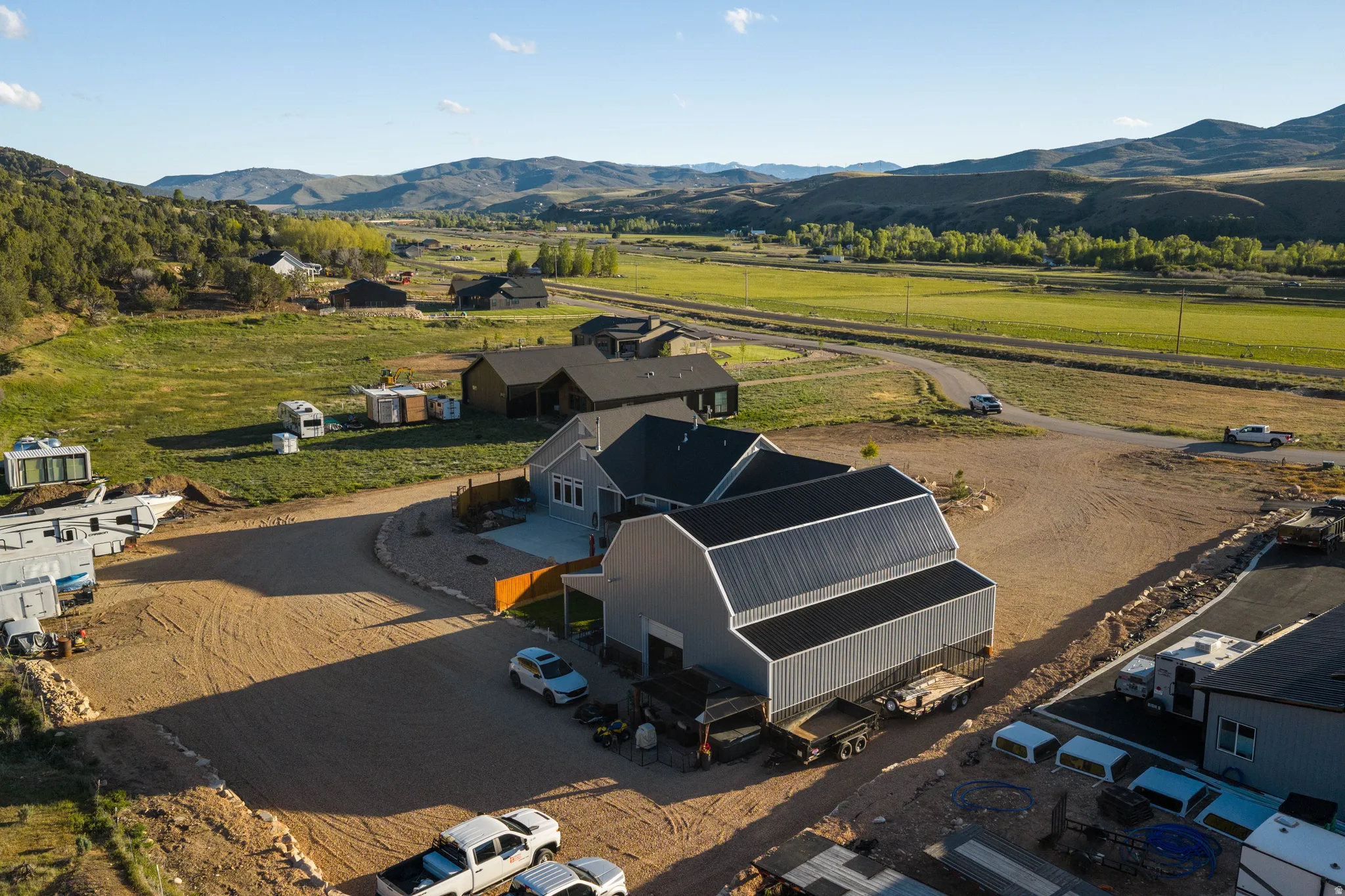Expansive property featuring a contemporary home with light grey siding, a substantial detached outbuilding with a metal roof, a gravel driveway, and surrounding natural landscapes including distant mountains and green fields
