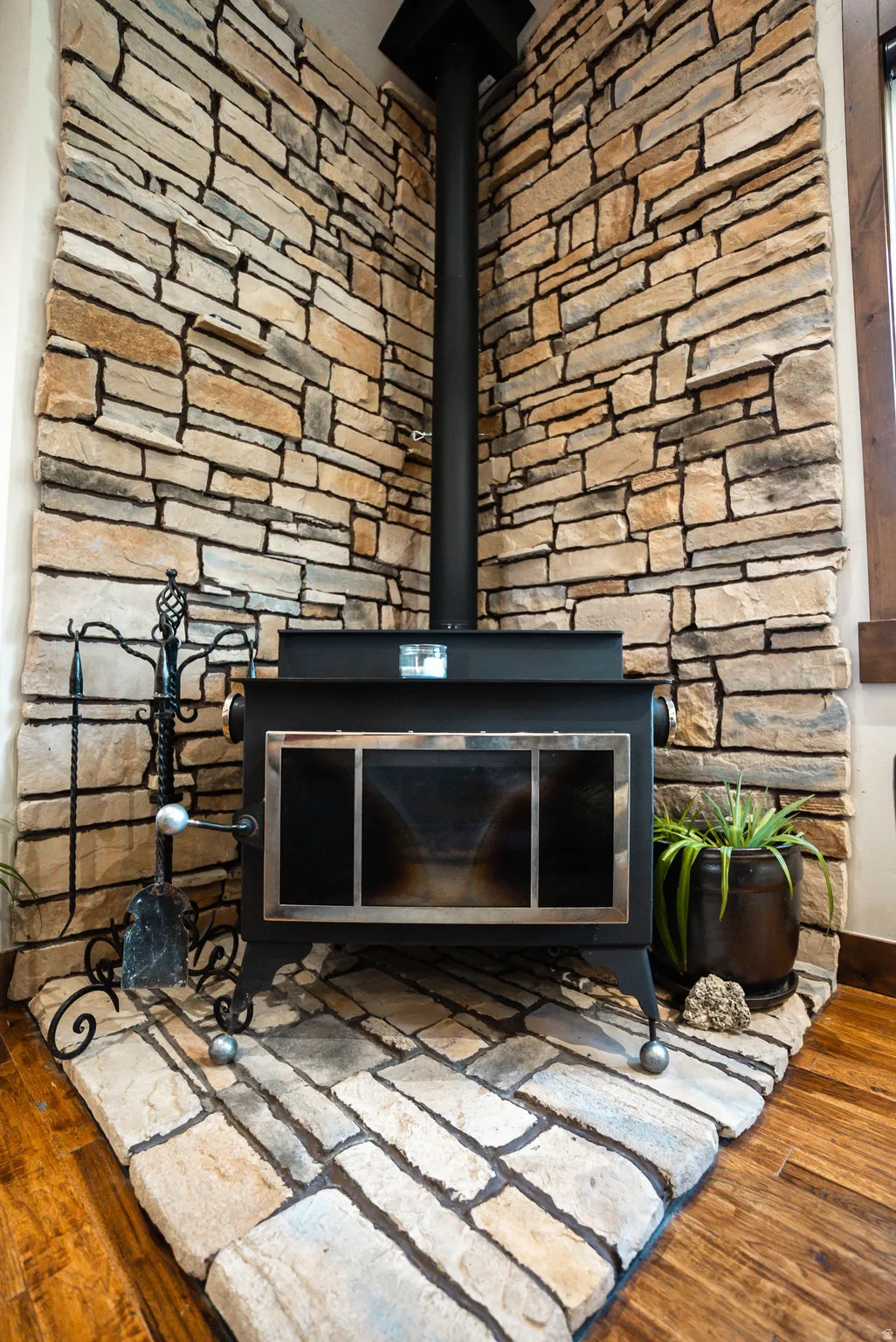 Corner wood-burning stove featuring a black stovepipe, stone surround, stone hearth, and wood-finish flooring