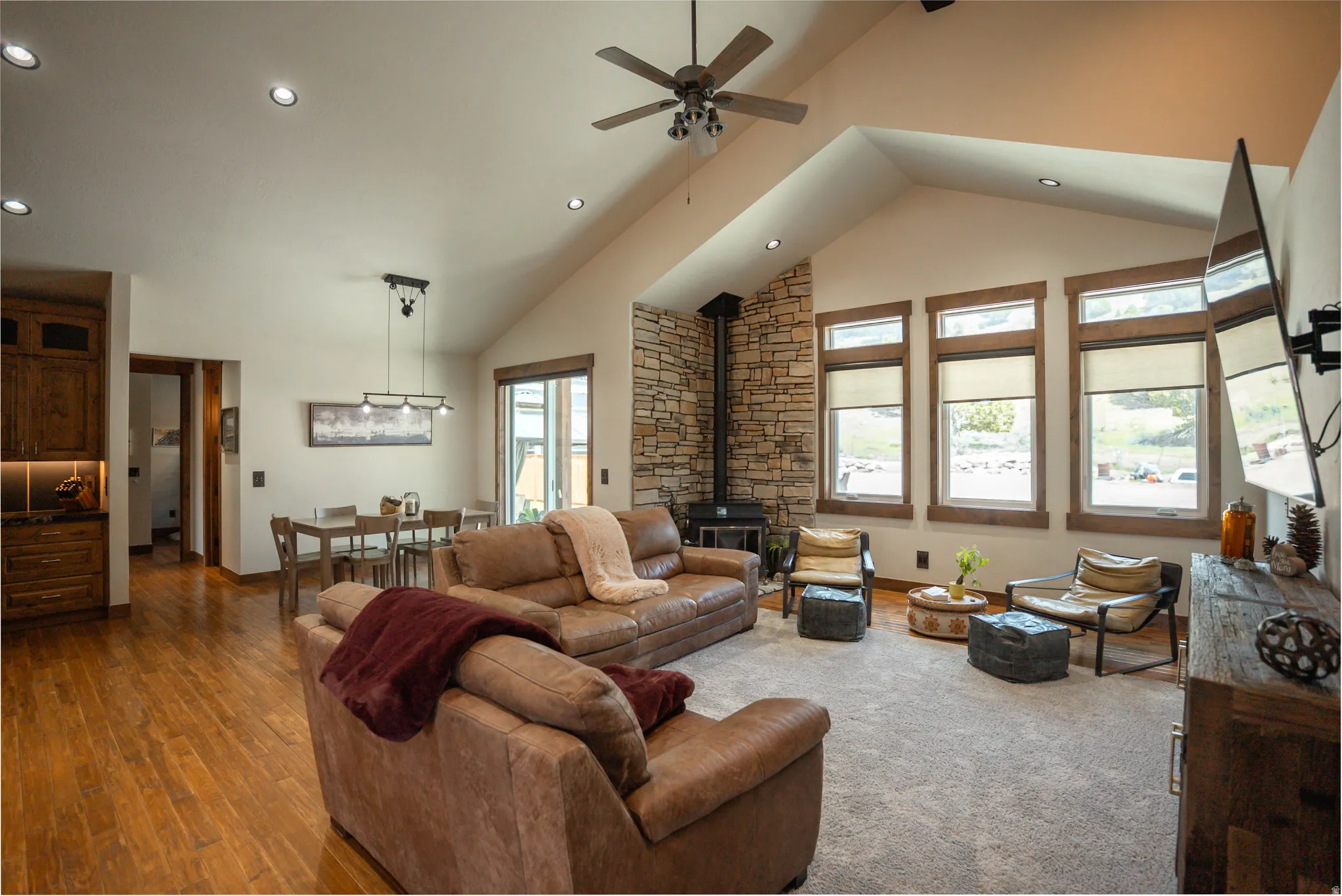 Vaulted ceilings with recessed lighting, stone-clad fireplace, wood-finish flooring, and multiple windows with roller shades