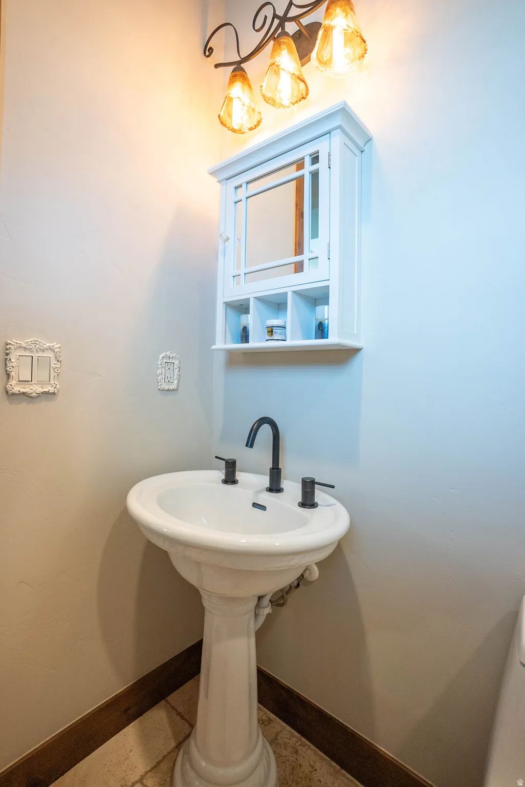 Pedestal sink with matte black fixtures, complemented by a white mirrored wall cabinet and an overhead three-bulb vanity light