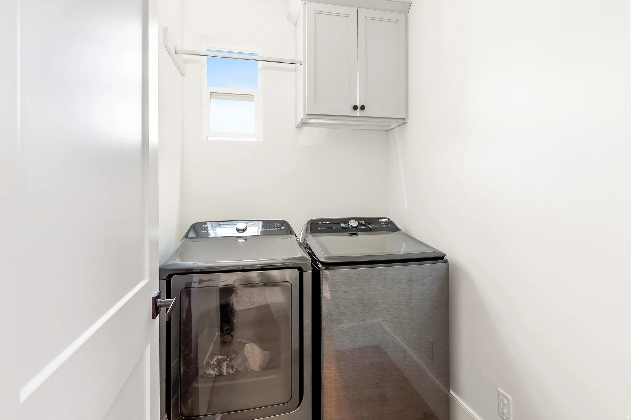 Laundry area featuring bright white walls, a small high window, white shaker-style cabinetry with dark hardware, and a chrome hanging rod