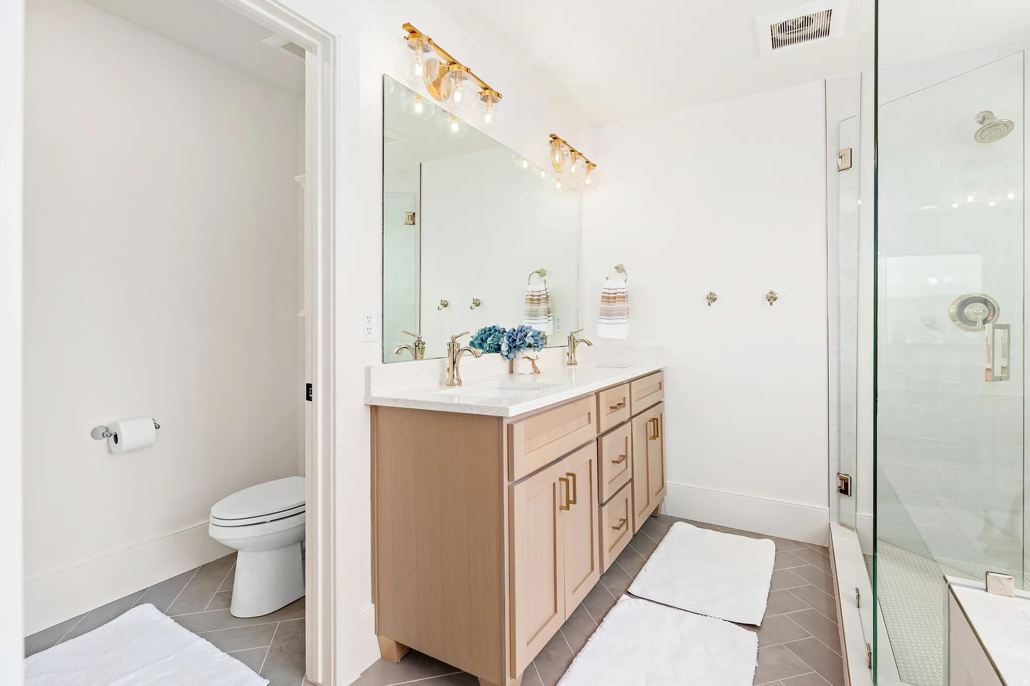 Bathroom featuring a dual vanity with a light wood-finish cabinet and stone countertop, a large framed mirror, and a glass-enclosed shower with a rainfall showerhead