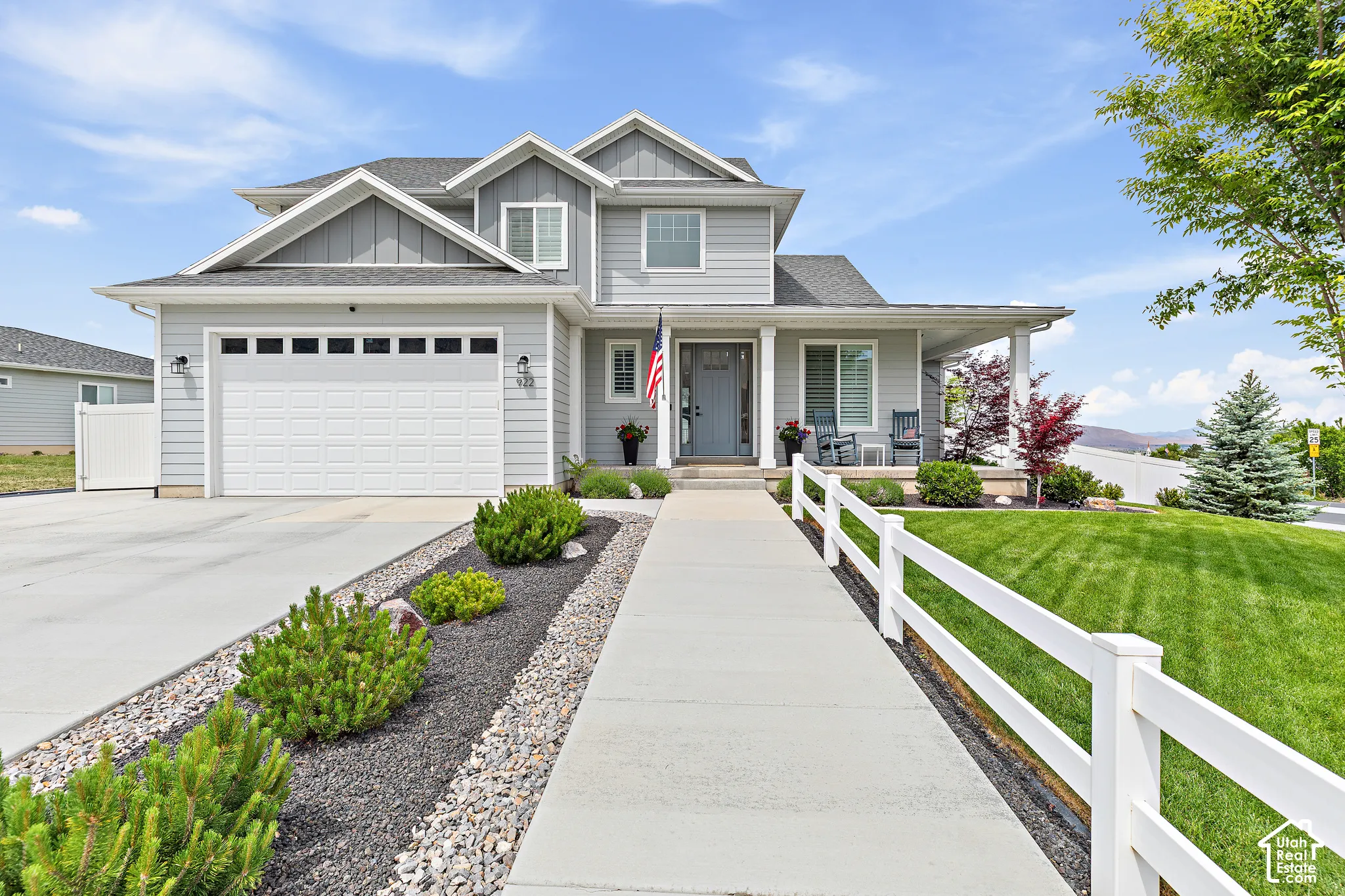 Two-story residence featuring light gray siding, a covered front porch, an attached two-car garage, a concrete driveway, and a white picket fence bordering a green lawn