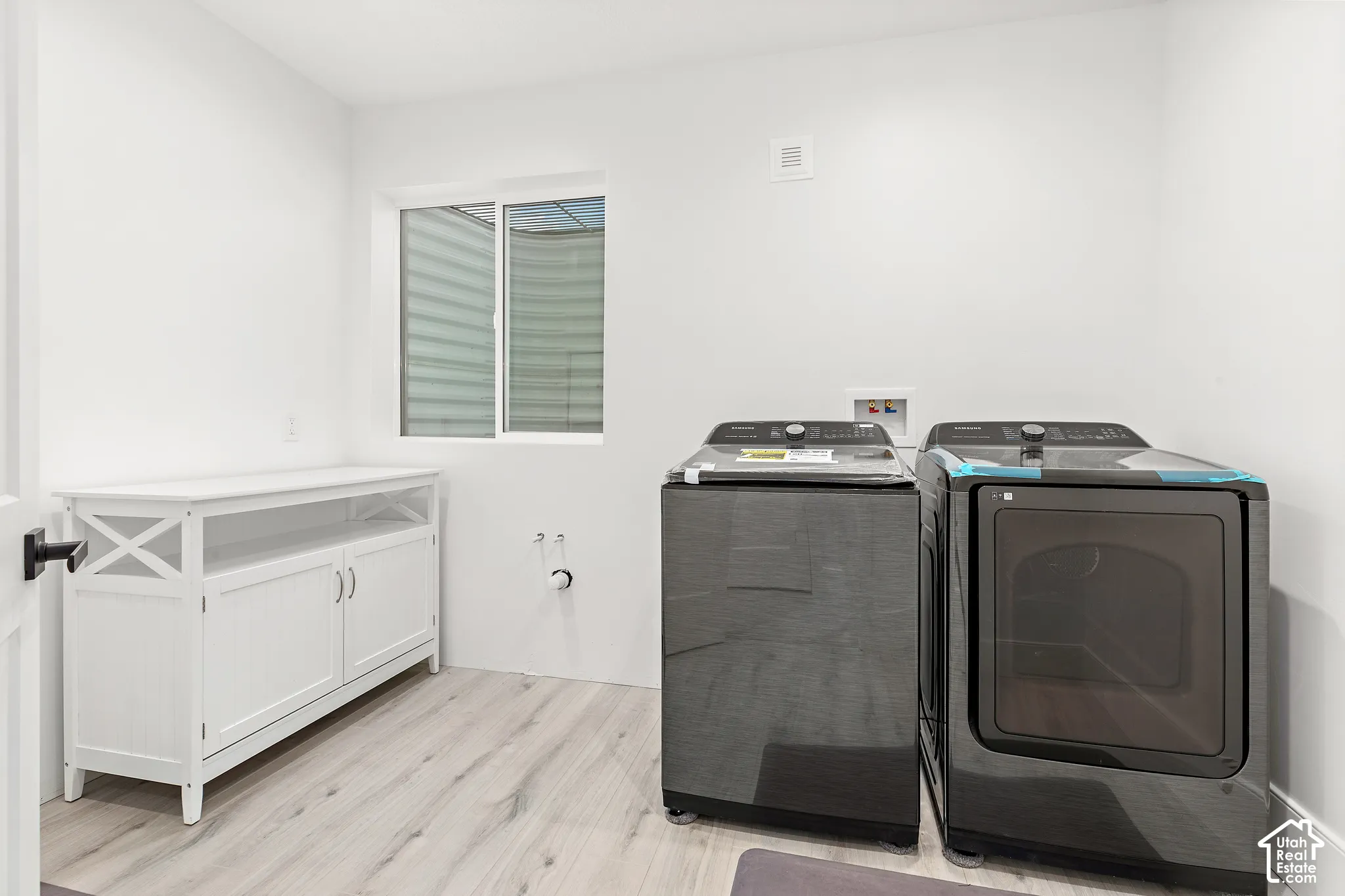 Laundry room featuring light wood-finish flooring, a white built-in storage cabinet, a window, and white walls