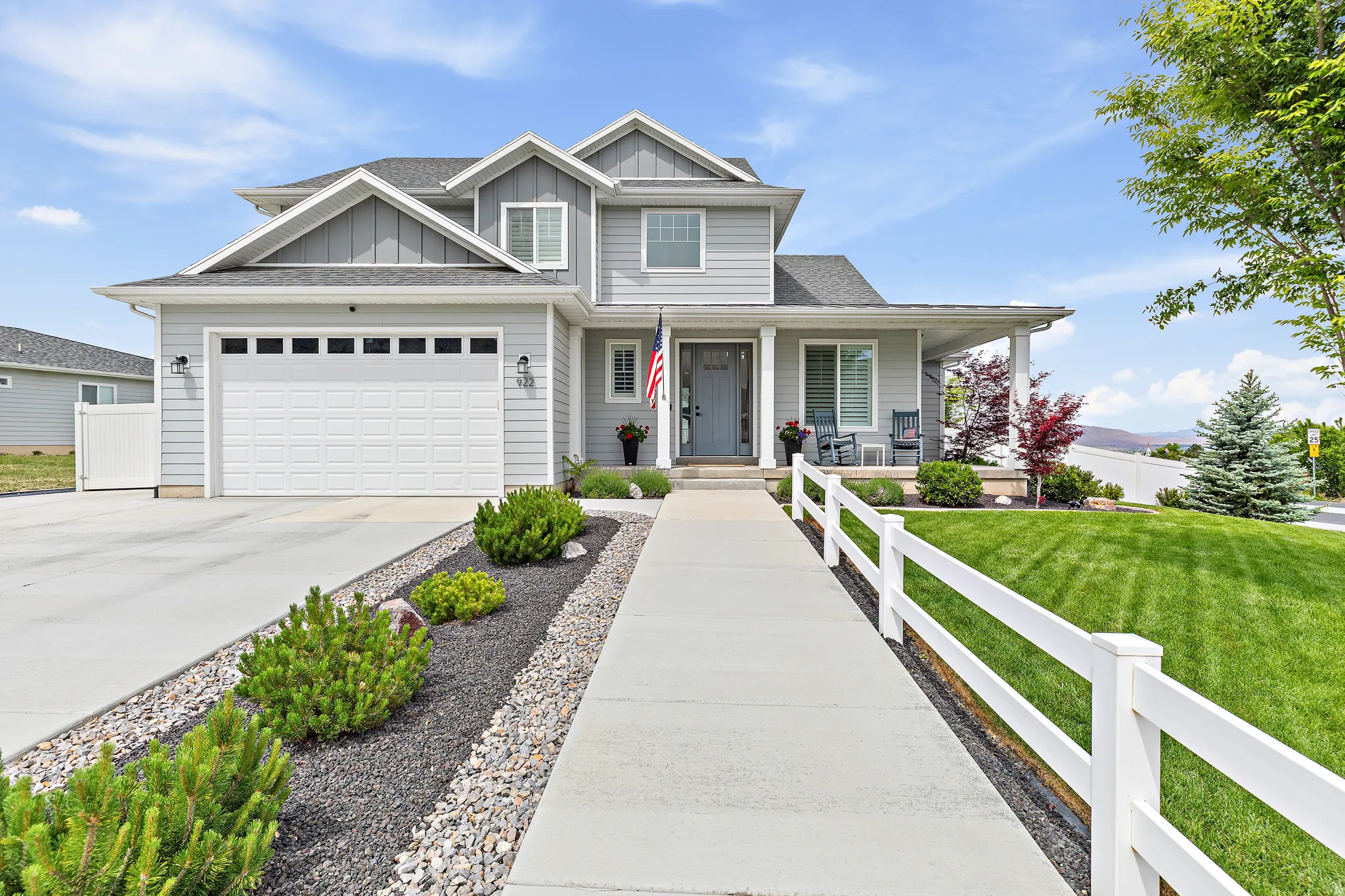 Two-story residence featuring light gray siding, a covered front porch, an attached two-car garage, a concrete driveway, and a white picket fence bordering a green lawn