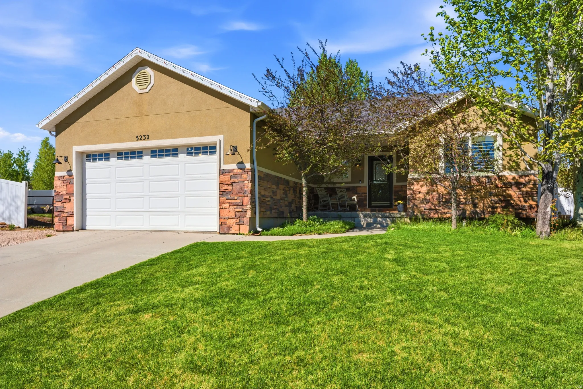 Exterior featuring tan stucco siding, stone accents, an attached garage, a paved driveway, and a front lawn with mature trees