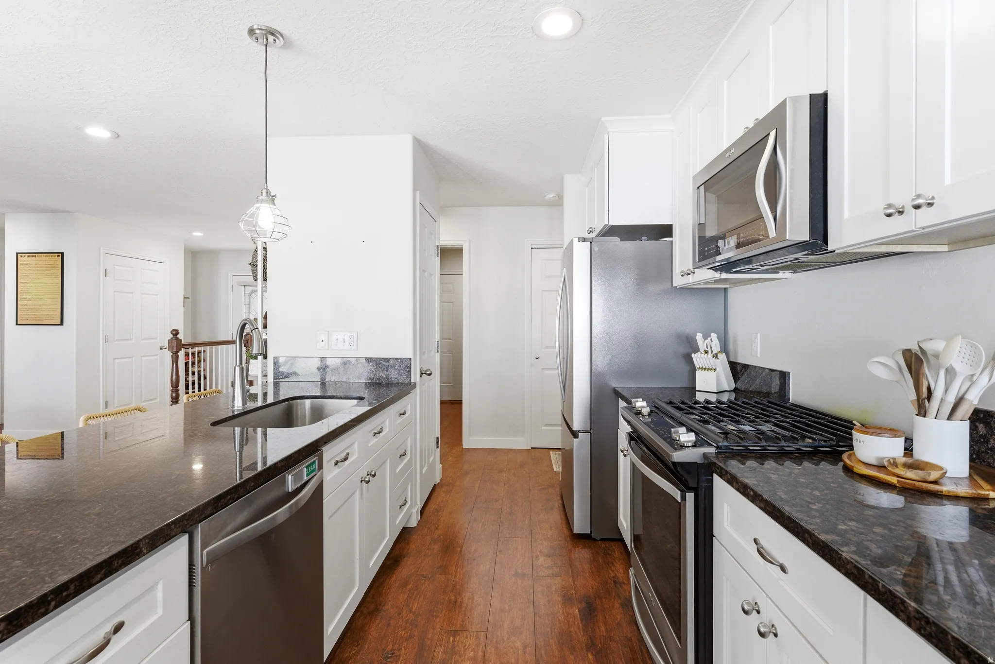 Modern kitchen featuring white cabinetry, dark stone countertops, stainless steel appliances, wood-finish flooring, and a central island with a sink and pendant lighting