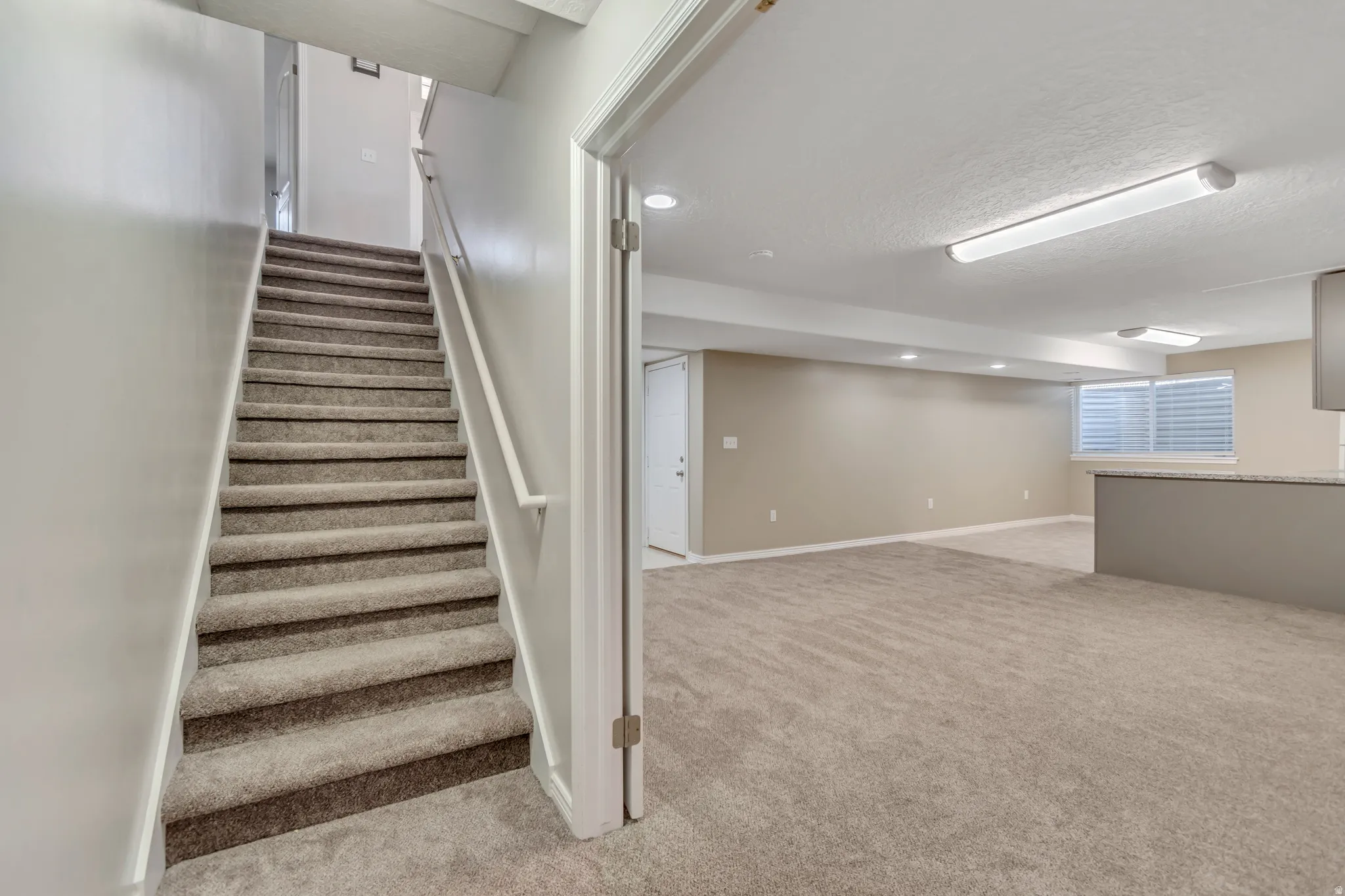 Carpeted staircase with white handrail leading to a spacious lower level featuring light beige walls and recessed lighting