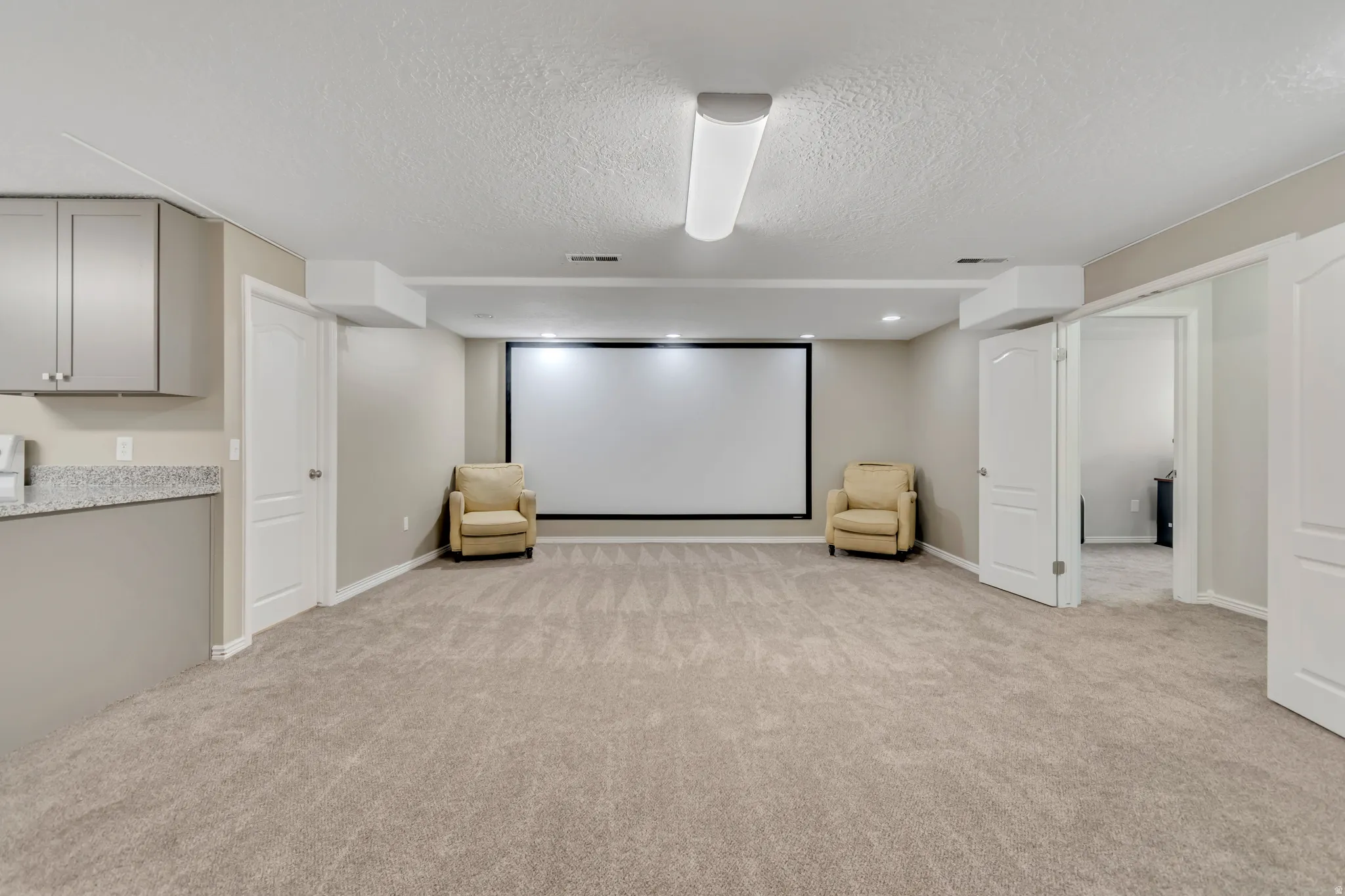 Spacious entertainment area with neutral-toned carpeting, a large projection screen, recessed lighting, and a built-in wet bar featuring gray cabinetry and a speckled countertop