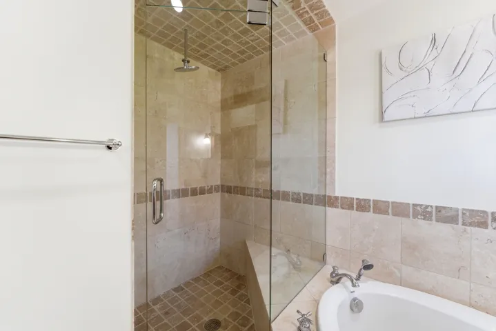 Glass-enclosed shower featuring a rainfall showerhead, built-in bench, and floor-to-ceiling tile work, complemented by a soaking tub with polished chrome fixtures and a natural stone tile surround