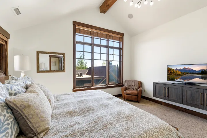 Vaulted ceiling with exposed wood beam, large multi-pane window with wood trim, wall-mounted media console, and recessed lighting fixtures