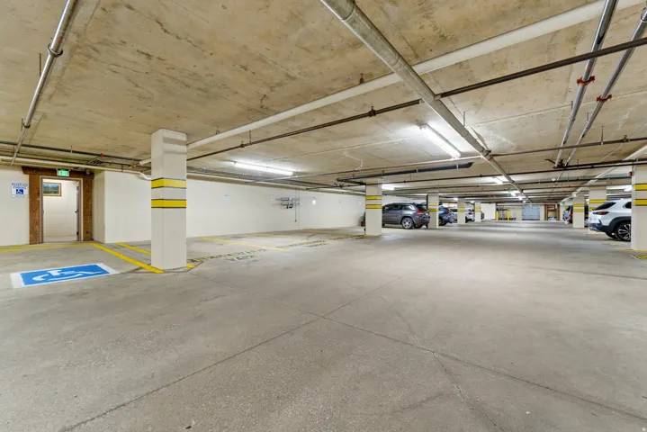 Spacious underground parking area featuring concrete flooring, exposed pipework, and fluorescent lighting