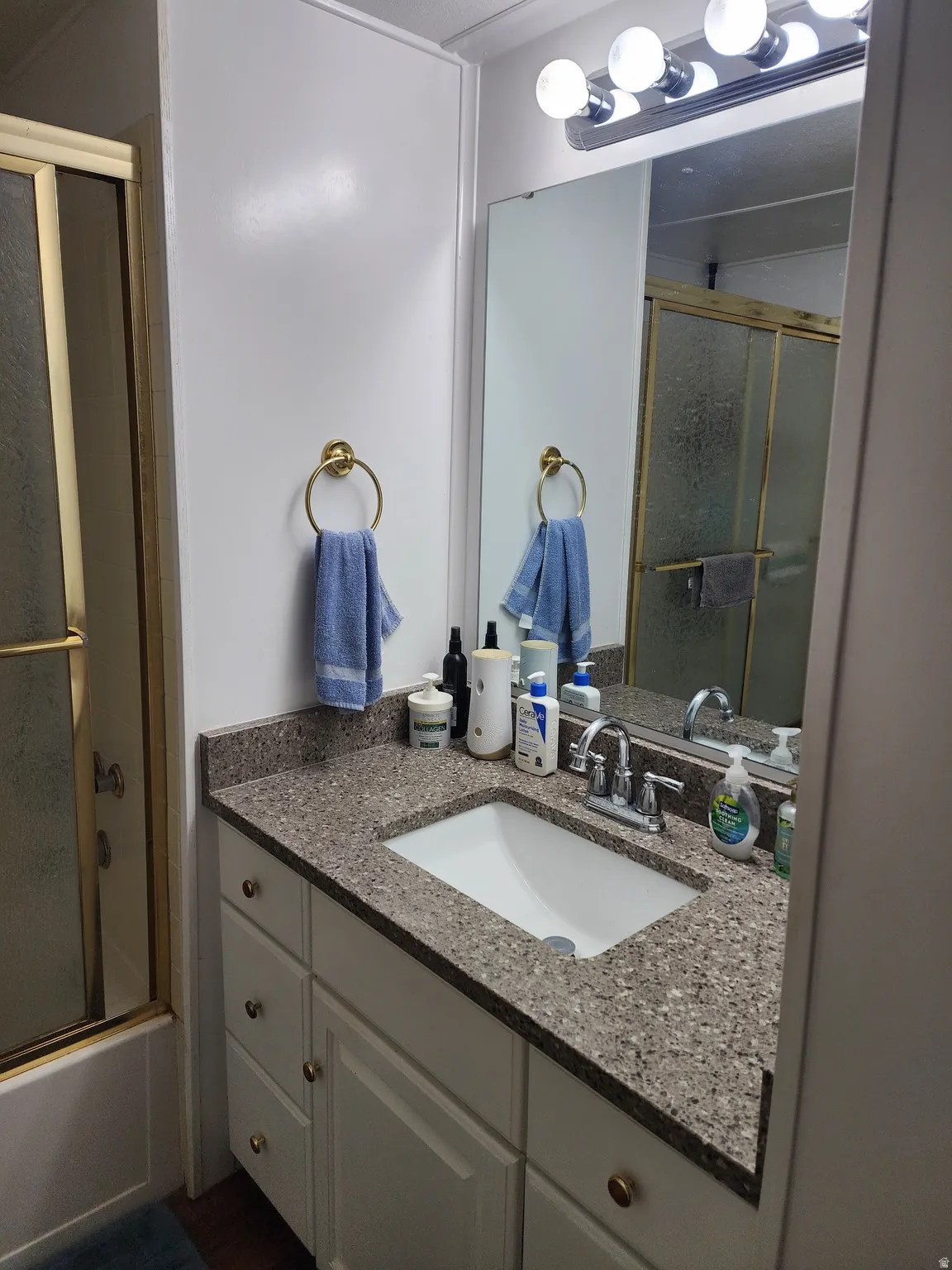 Bathroom featuring a white vanity with gold-toned hardware, a speckled countertop, an integrated sink, and a wall-mounted mirror with vanity lighting