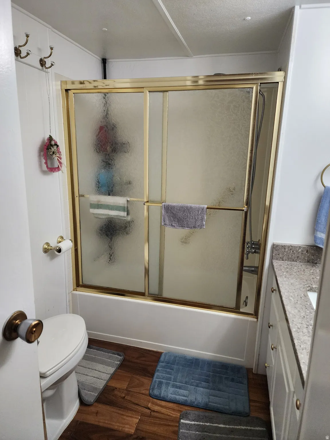 Bathroom featuring a tub/shower combination with gold-tone framed sliding doors, a vanity with a speckled countertop, and wood-finish flooring