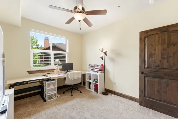 Carpeted room featuring a large window with white plantation shutters, a dark wood-finish door, matching wood-finish baseboards, and a ceiling fan with integrated lighting