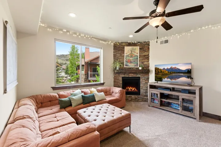 Living area featuring a stone accent wall with a gas fireplace, a large window, recessed lighting, a ceiling fan with integrated lighting, and neutral-toned carpet flooring